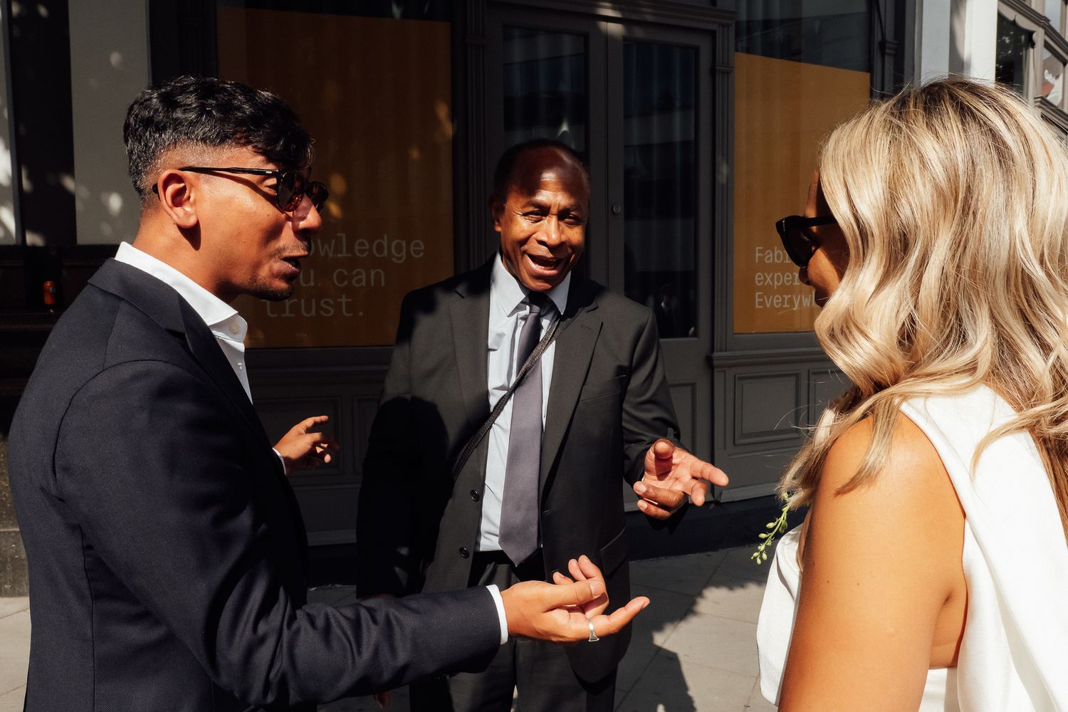 Two men in dark suits gesture while talking outdoors with a blonde woman in a white sleeveless top and sunglasses in the foreground.