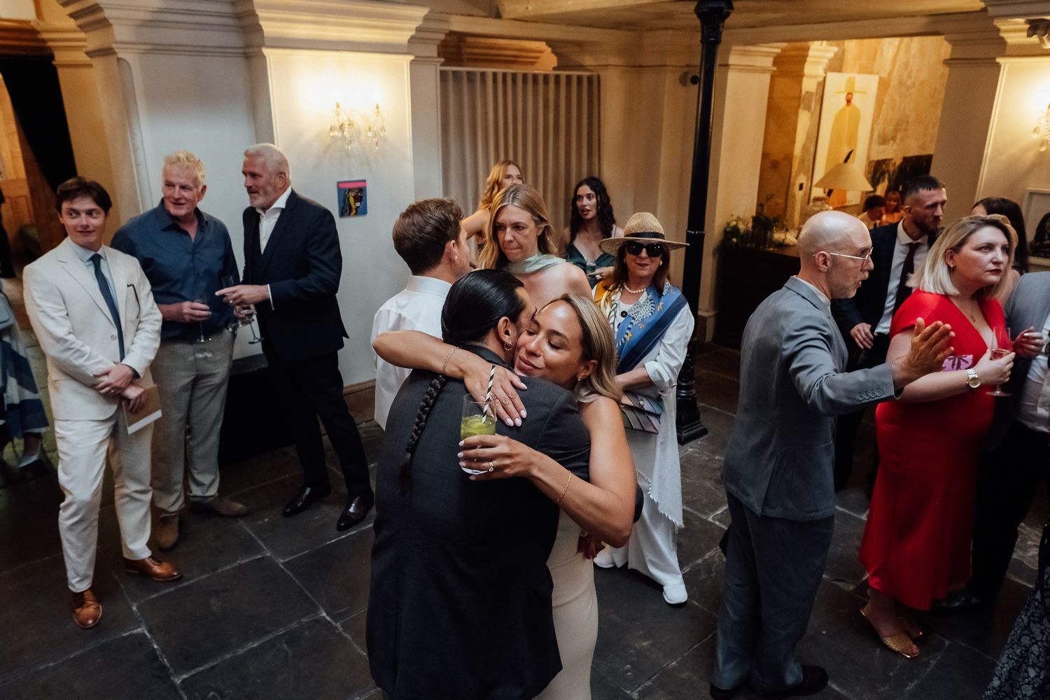 Wedding guests in formal attire embrace and mingle on a stone-tiled floor inside an elegant historic venue with arched doorways.