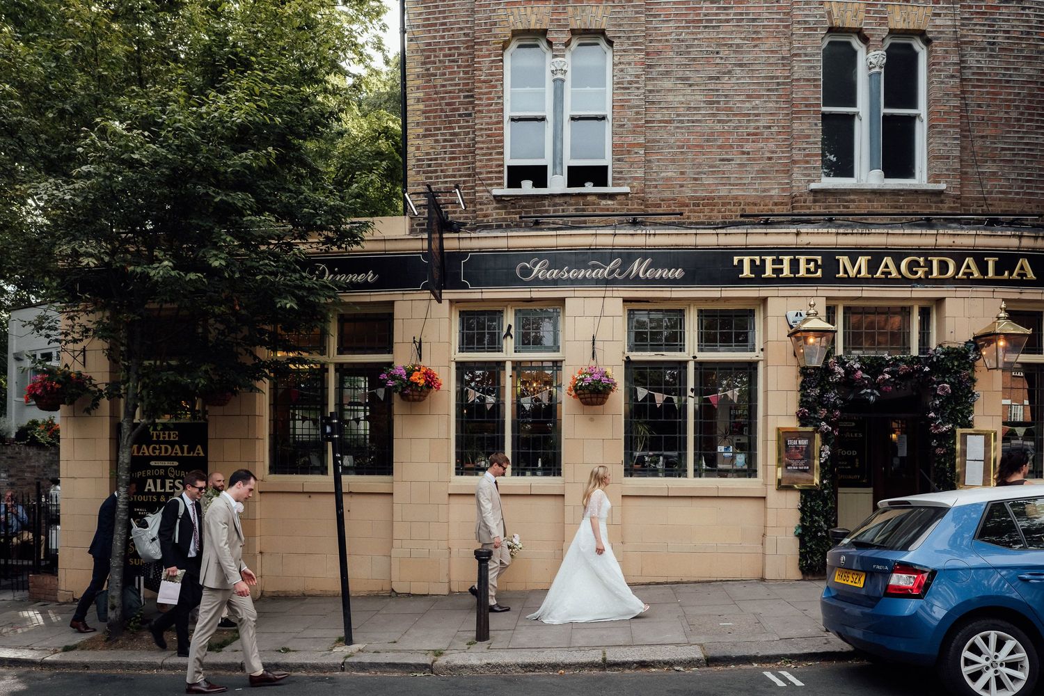Bride in a white wedding dress walks along the pavement outside The Magdala pub, a brick London building, as guests in suits look on.