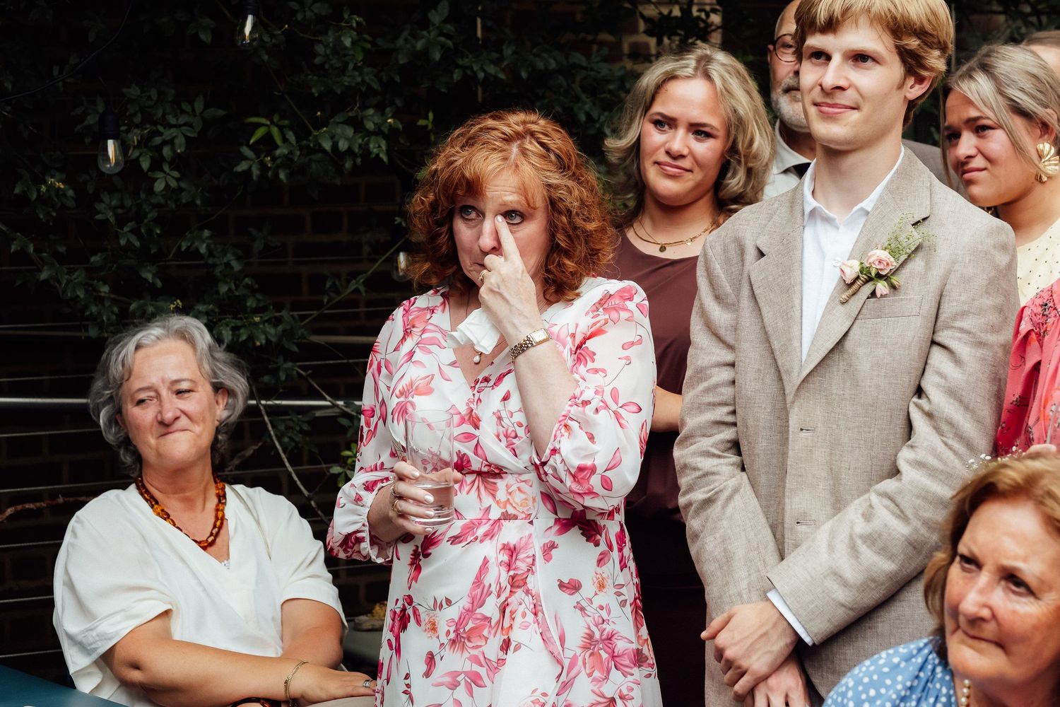 A red-haired woman in a floral dress wipes away tears at an outdoor wedding as guests in formal attire stand and sit around her.
