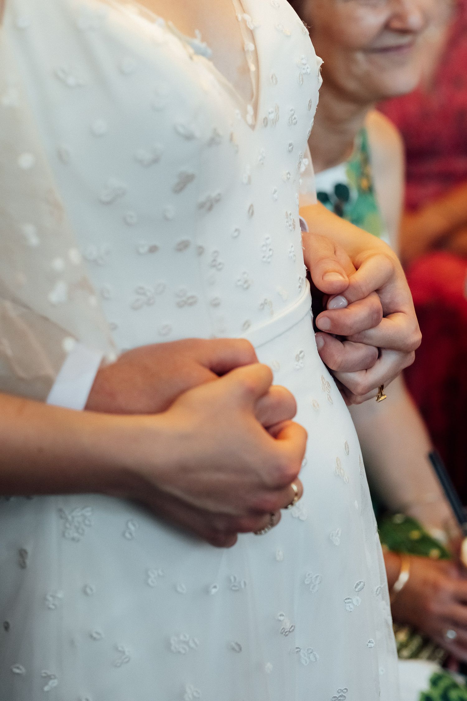Close-up of a bride in a white lace wedding dress holding hands with her partner as a smiling guest looks on.