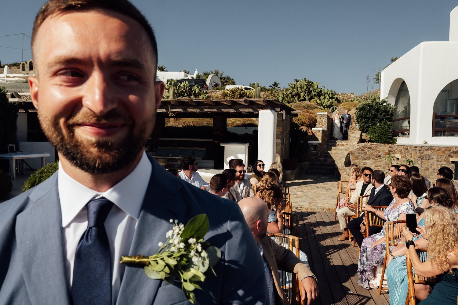 Smiling groom in blue suit with boutonniere at outdoor wedding ceremony in Mediterranean setting.