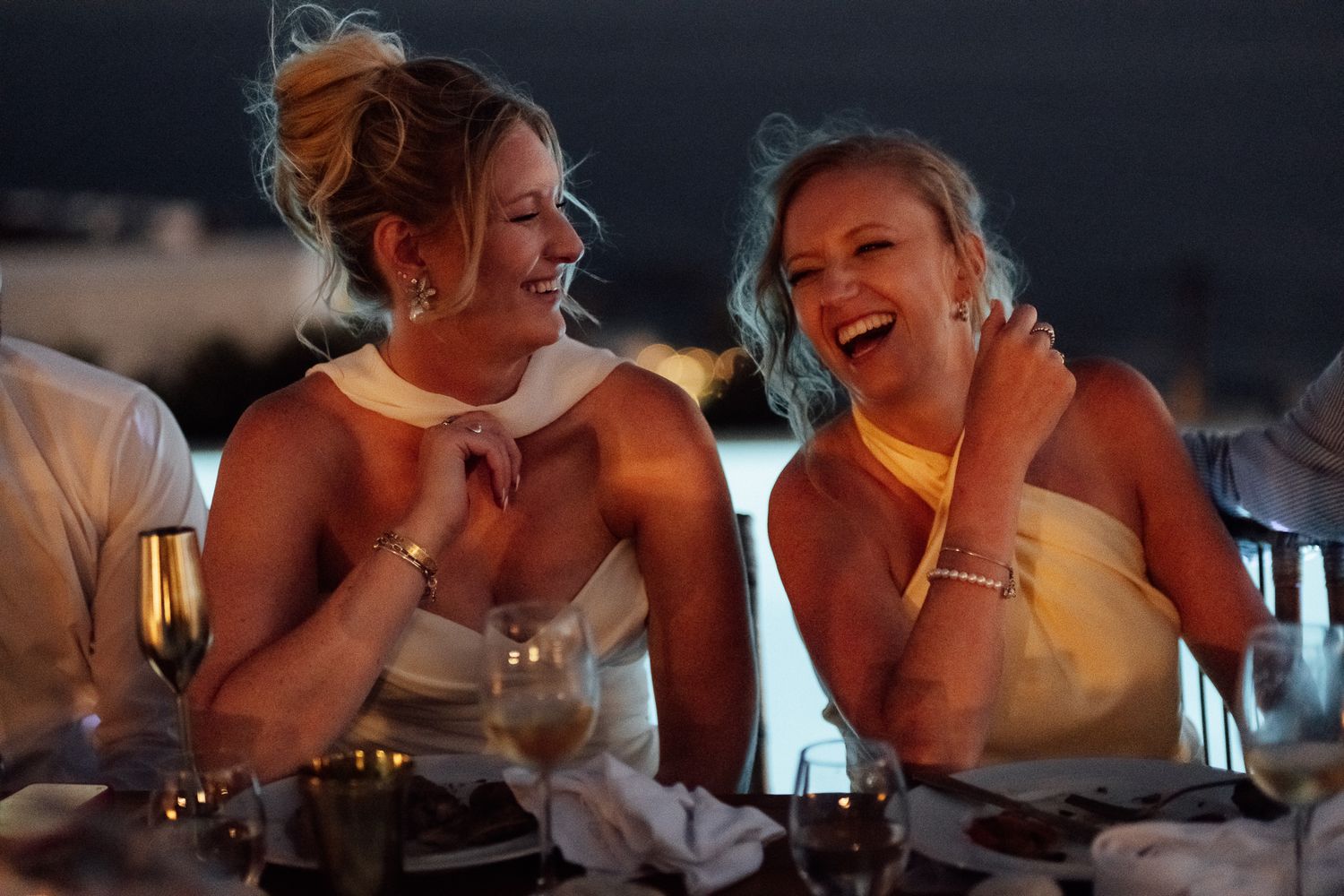 Two women in elegant dresses laughing joyfully at a formal dinner event with champagne glasses on the table.