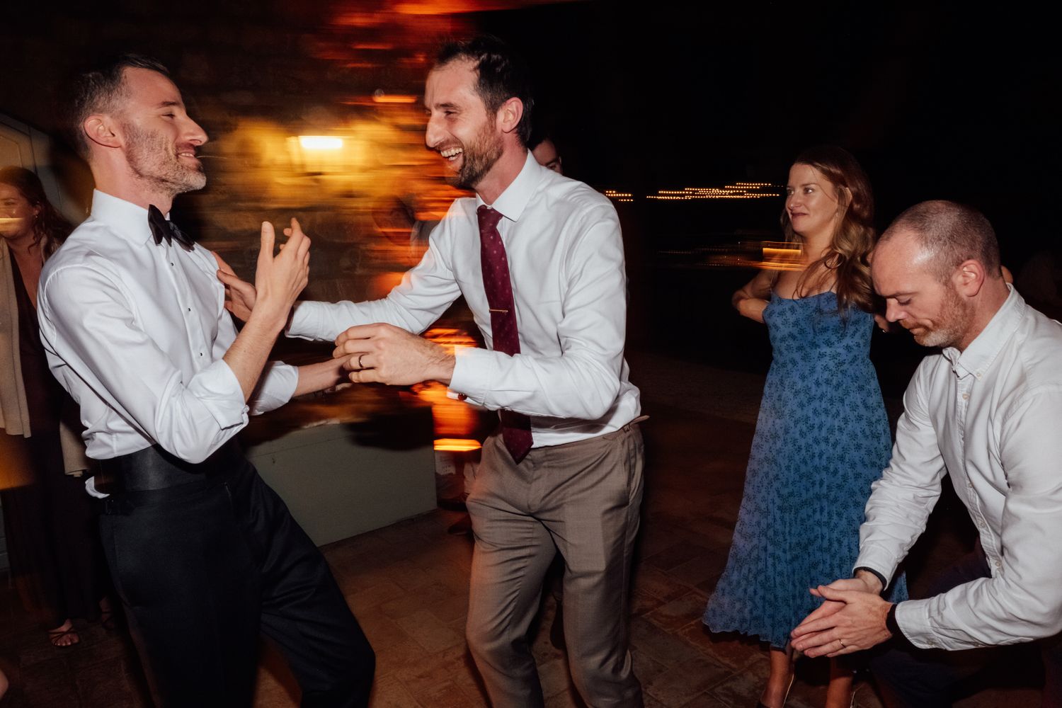 Two smiling men in dress shirts and ties dance joyfully together at a lively evening wedding reception as guests celebrate around them.