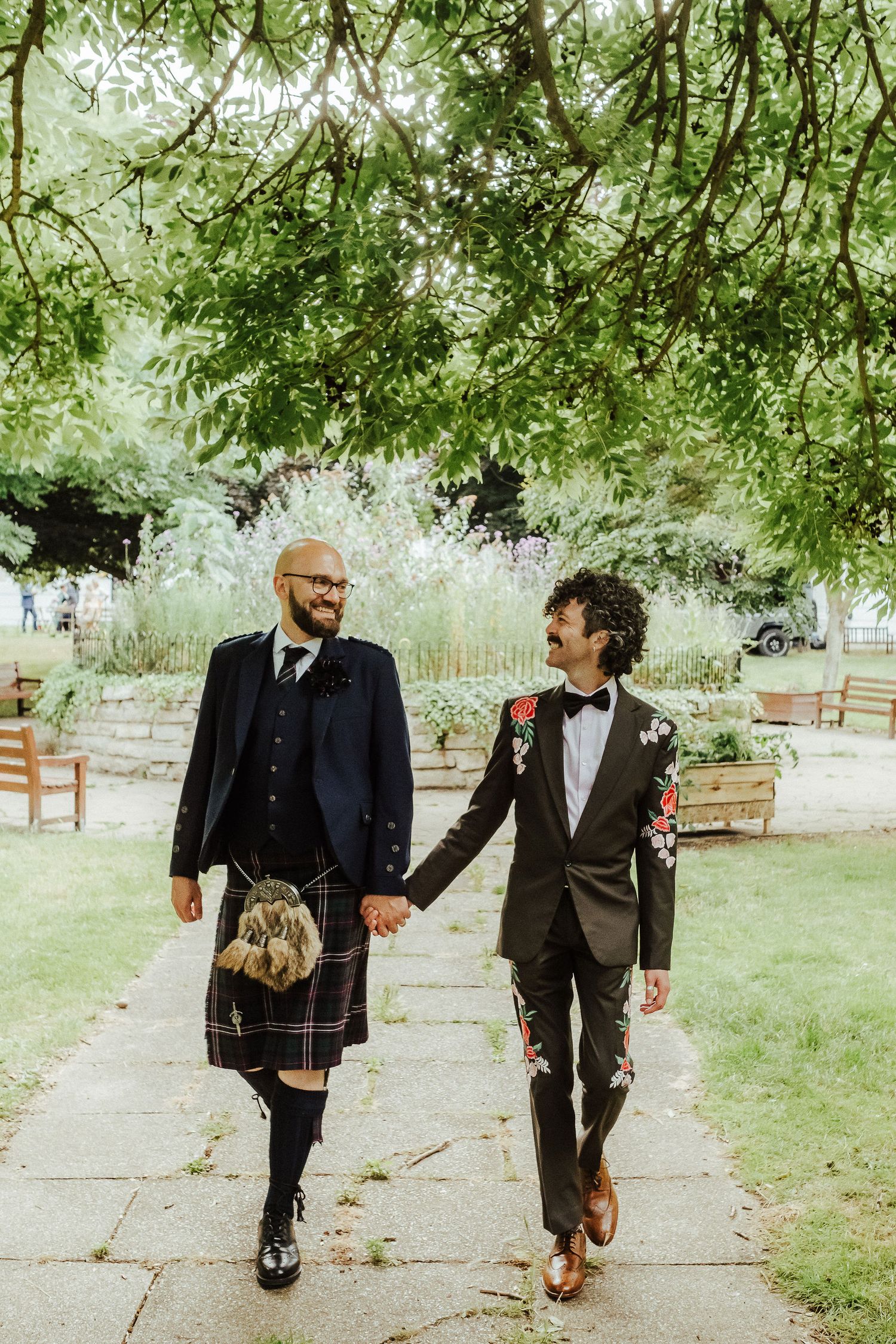 Two grooms walk hand in hand along a garden path under leafy trees, one in a traditional kilt outfit and one in a floral embroidered suit.