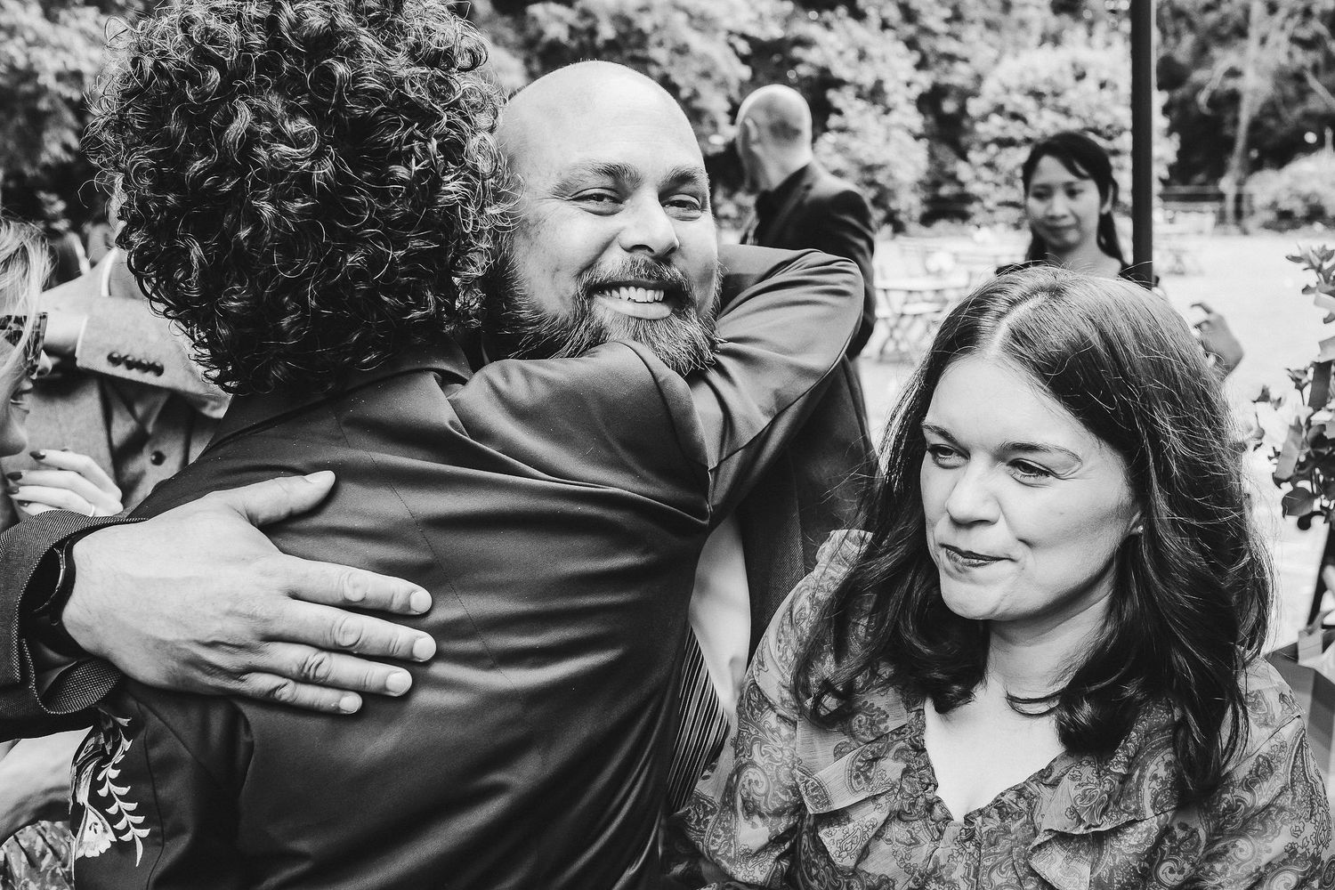 Black and white wedding photo of a smiling bearded man hugging a curly-haired guest while a woman watches nearby in a garden.
