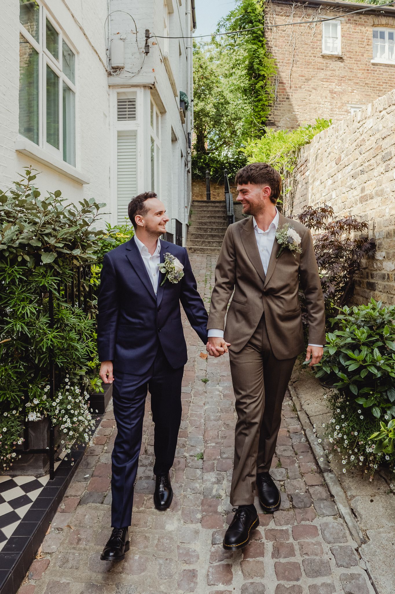 Two people in suits walking together and sharing a joyful moment on a brick pathway surrounded by greenery.