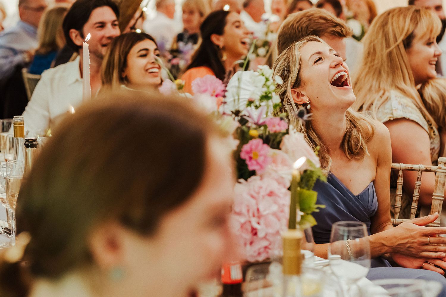 Wedding reception guests laugh heartily among pink floral centerpieces and candlelit tables.
