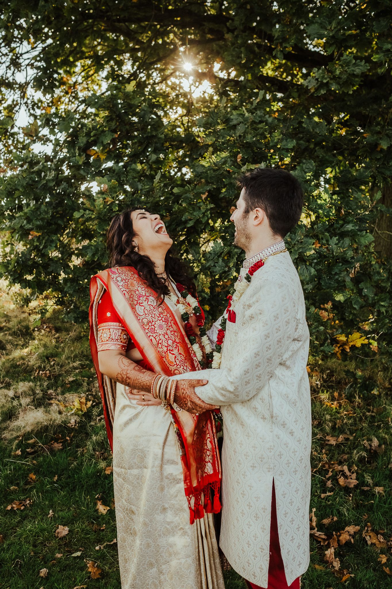 Joyful couple in traditional Indian wedding attire sharing a romantic moment under sunlit trees.