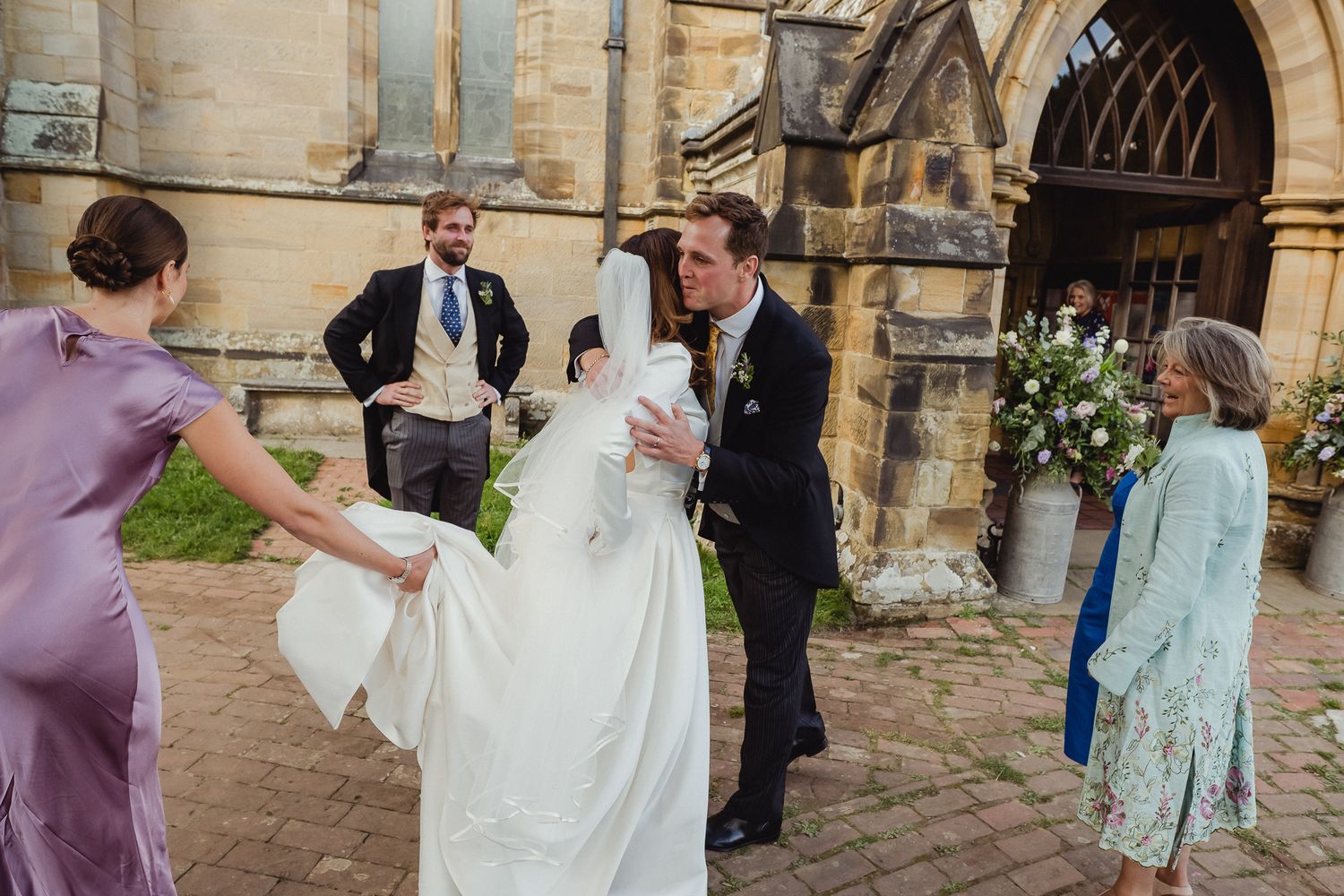 Wedding party helps with the bride's dress outside a historic stone church entrance adorned with floral decorations.
