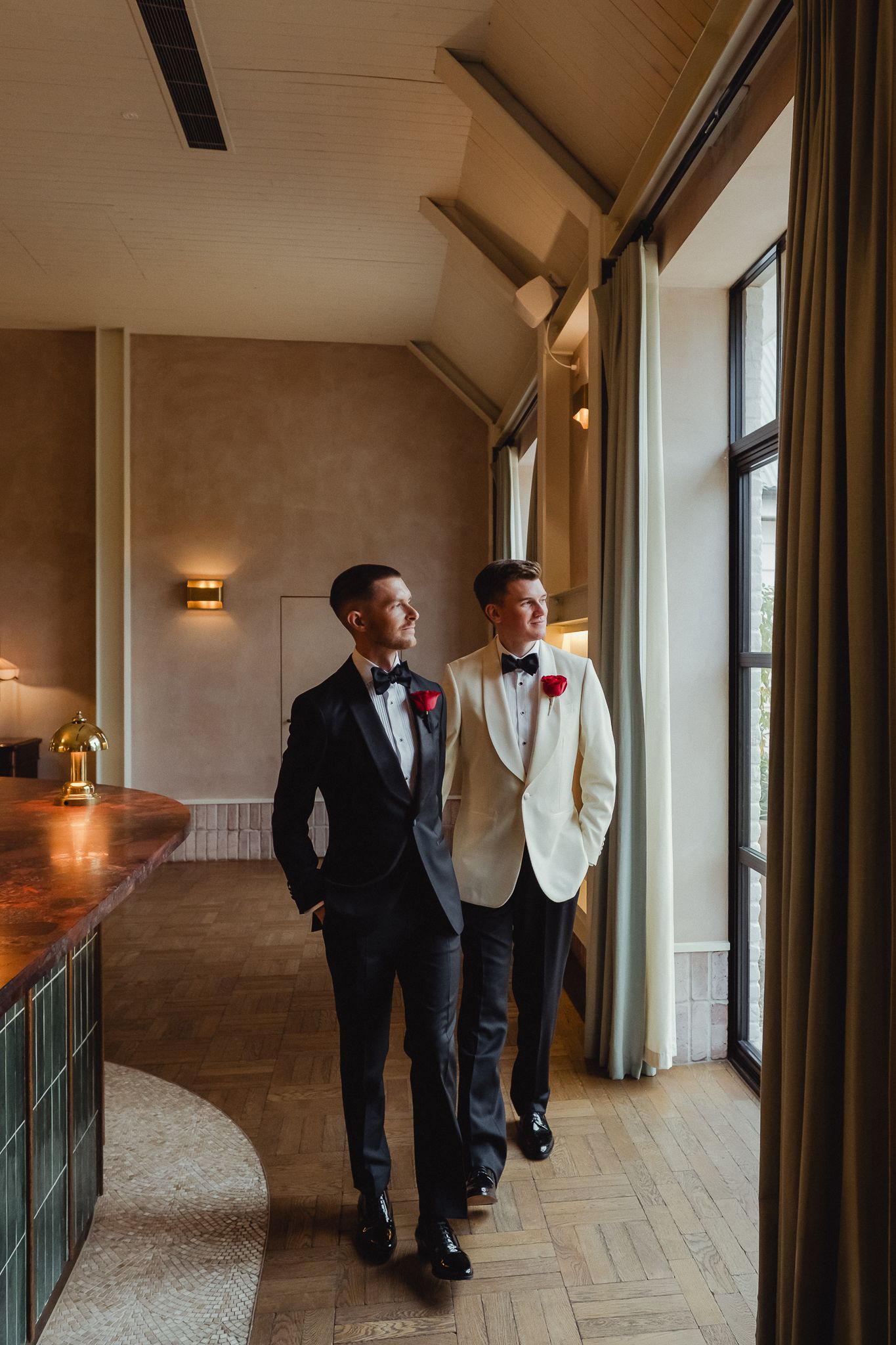 Groomsmen in formal attire walk down a modern hallway with large windows and wall sconces.