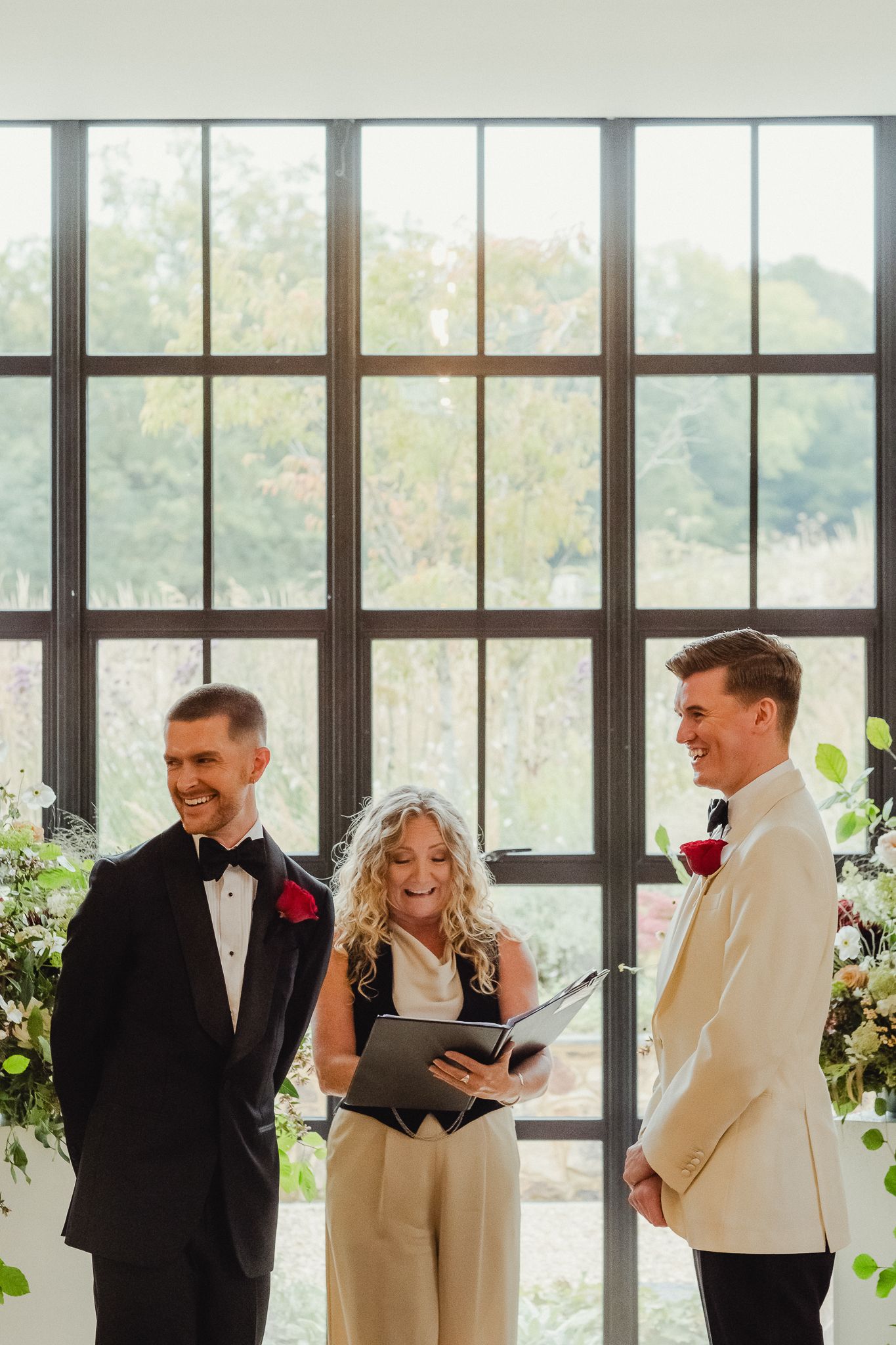 Wedding party members share laughter during an indoor ceremony by large windows with natural light streaming in.