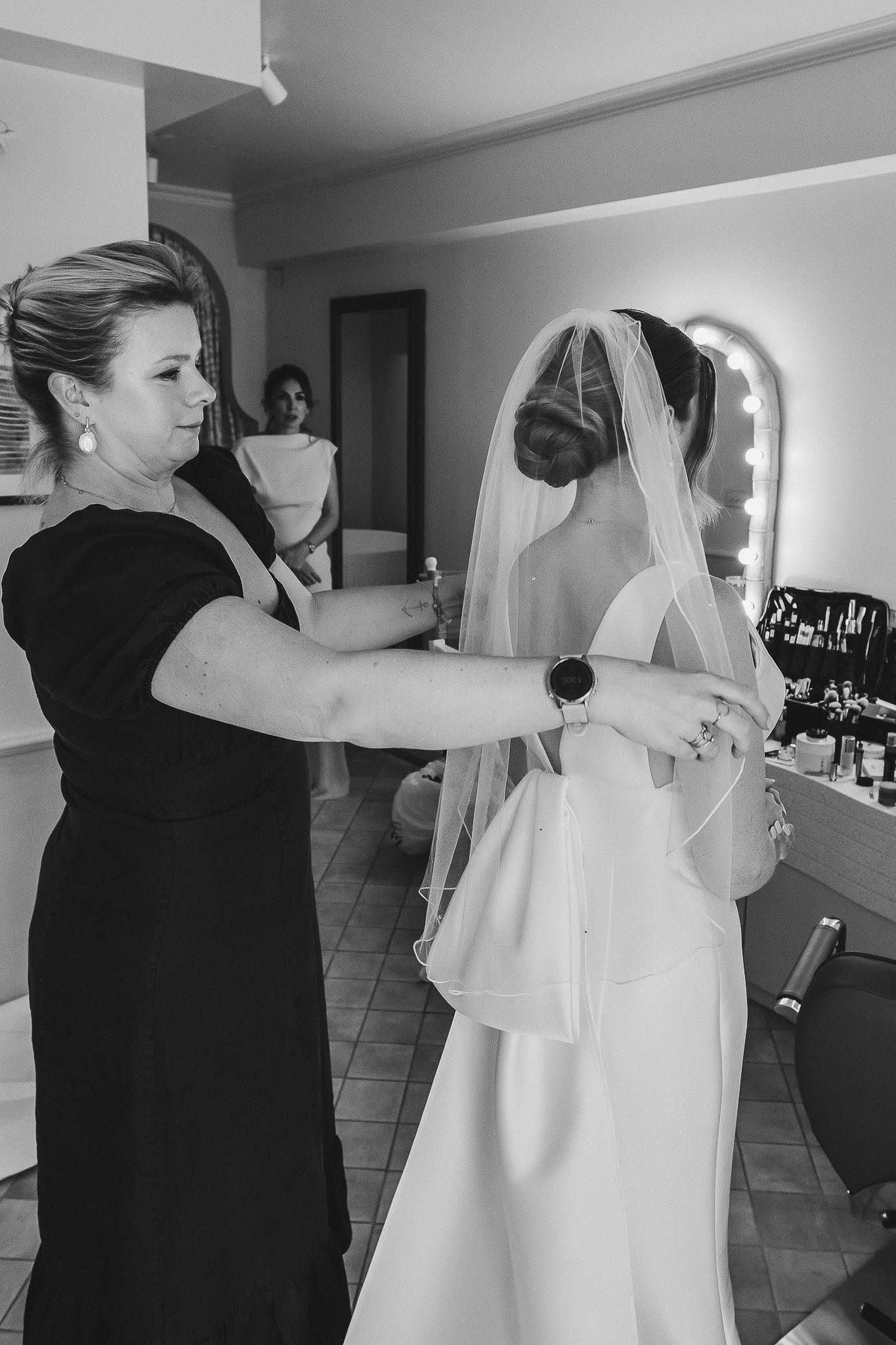 A bride getting ready on her wedding day as someone helps adjust her veil in a dressing room.