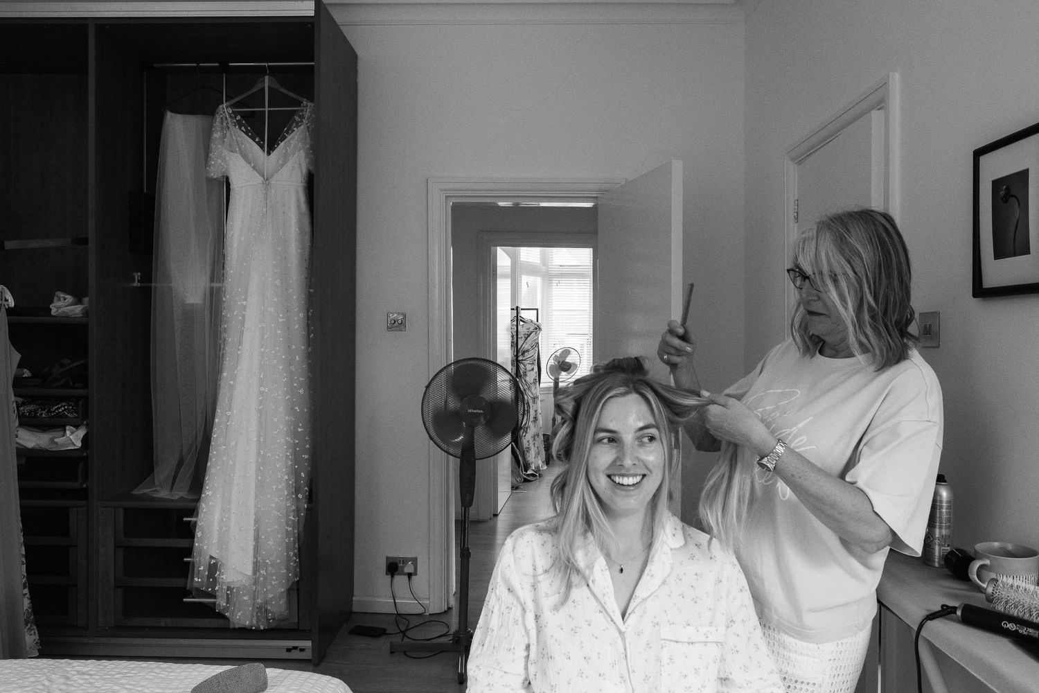 Smiling bride in a button-up shirt gets her hair styled by a hairdresser while a wedding dress hangs from a wardrobe in the background.