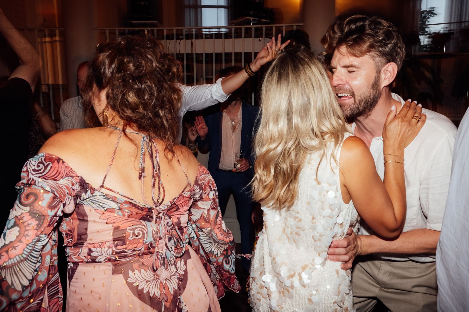 Wedding guests dance and embrace on a crowded dance floor, a woman in a floral dress with curly hair turned away from the camera.