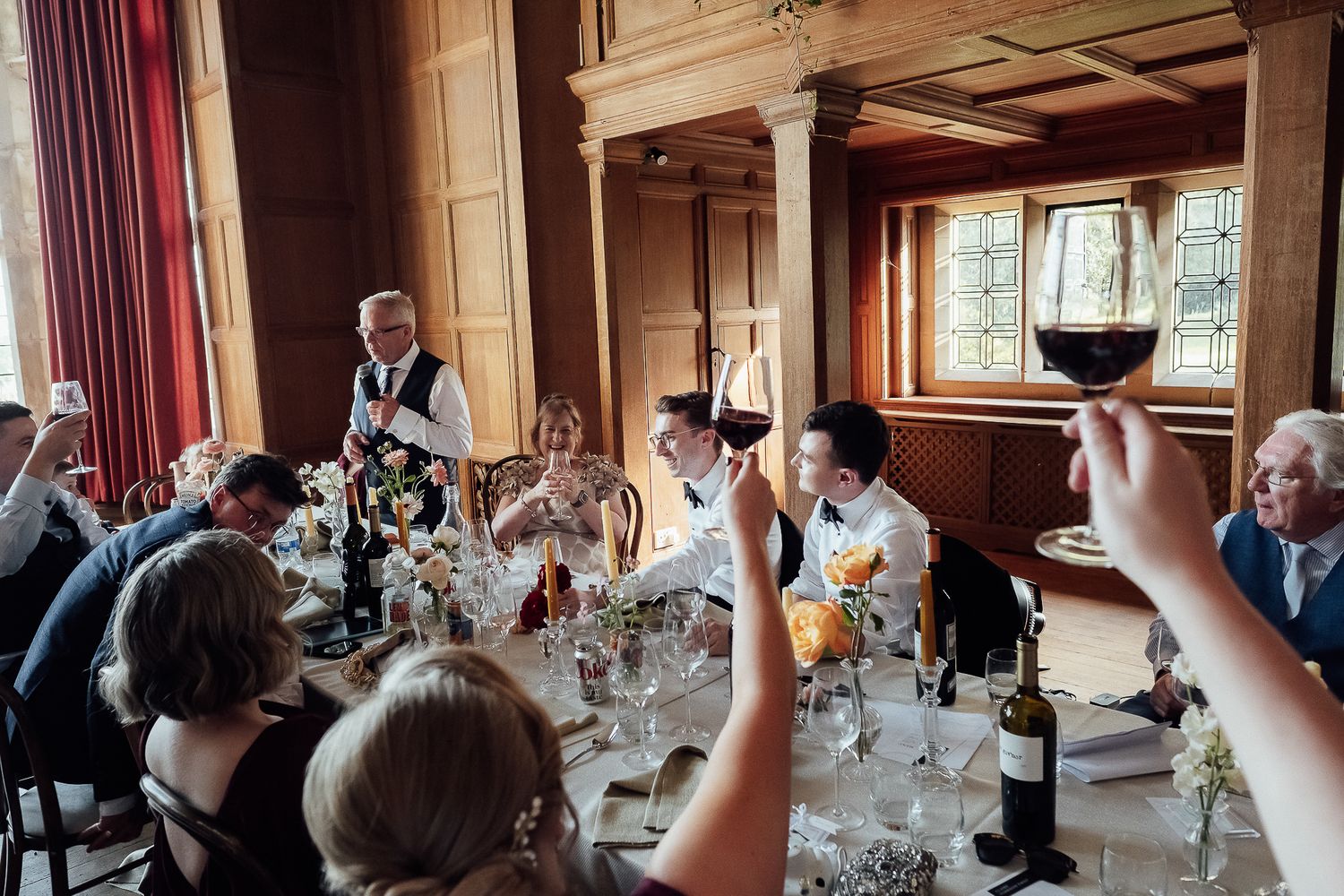 Elegant wedding reception toast in a wood-paneled hall, guests raising red wine glasses around a decorated dinner table.