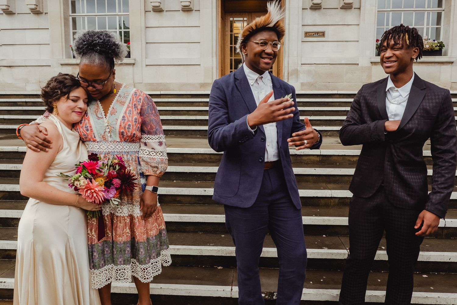 A wedding party laughs and smiles together while standing in front of a grand white building entrance.
