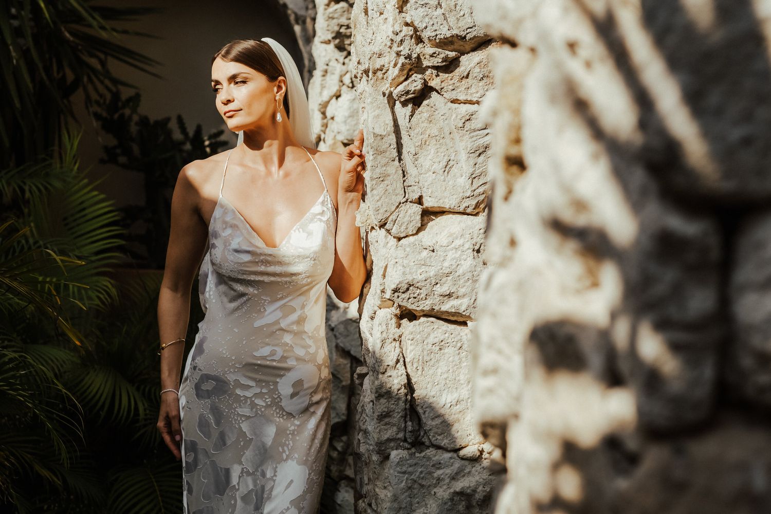 A bride in a silky wedding dress poses against a textured stone wall in dappled sunlight.