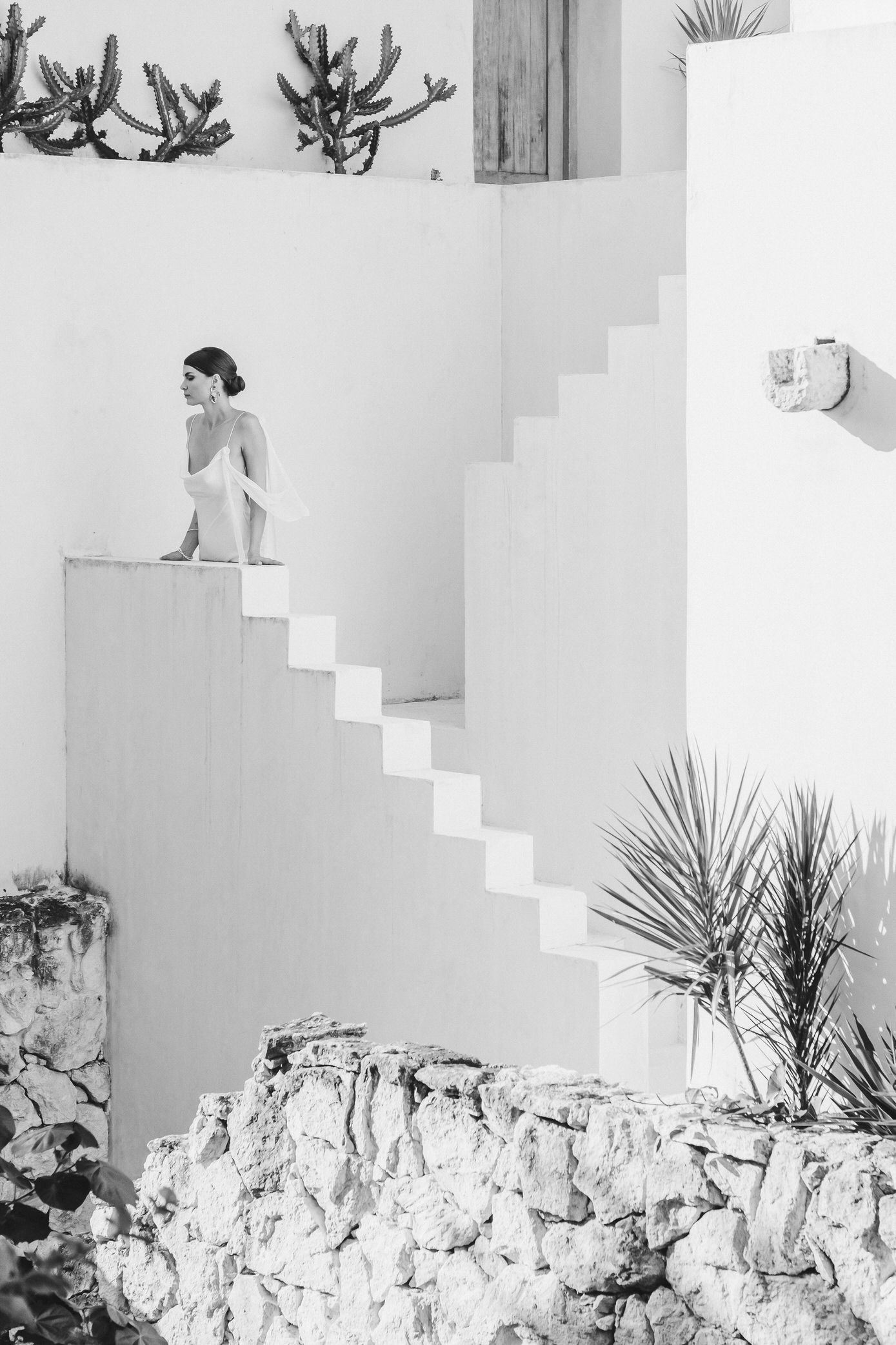A figure in a flowing white dress poses elegantly on white architectural steps with a stone wall backdrop.
