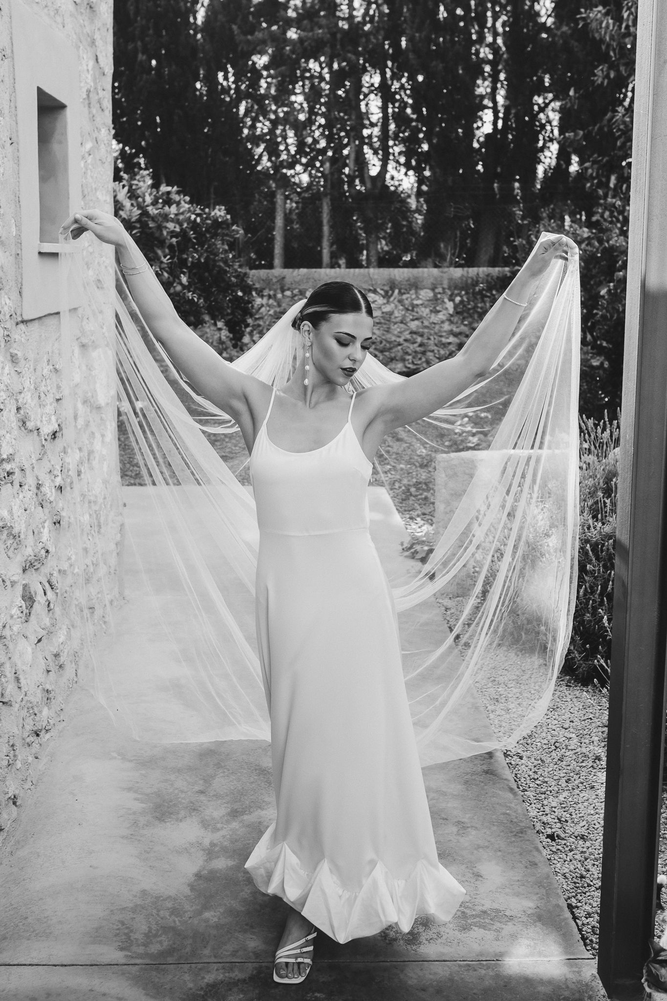 Black and white photo of a bride in a simple white gown holding up a flowing veil against a garden backdrop.