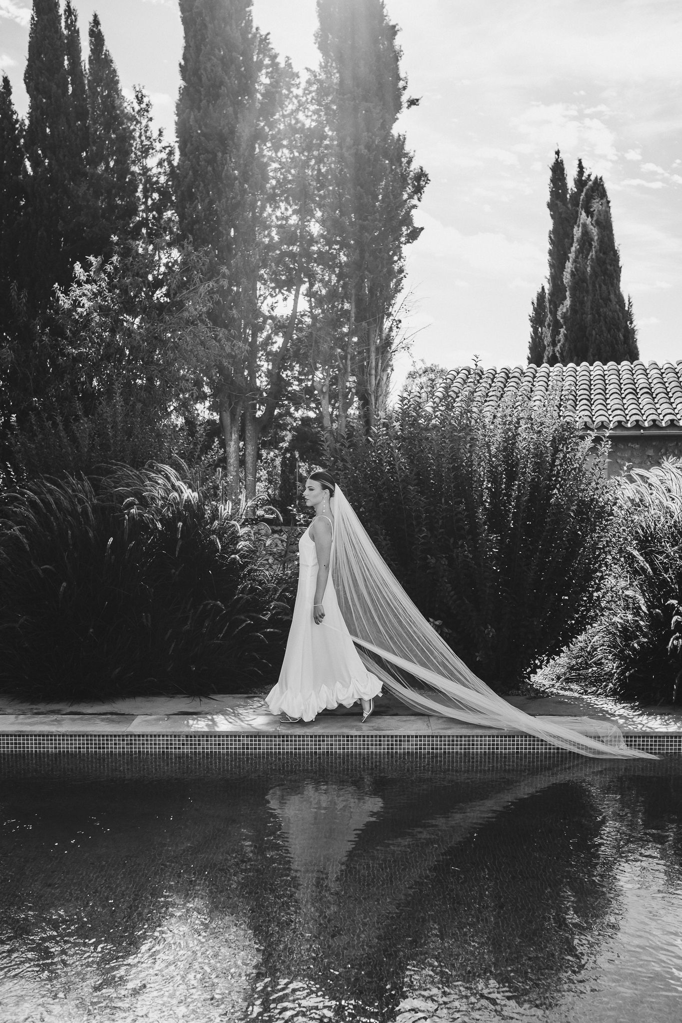 A flowing white wedding dress and veil create an ethereal reflection in a tranquil pond surrounded by cypress trees.