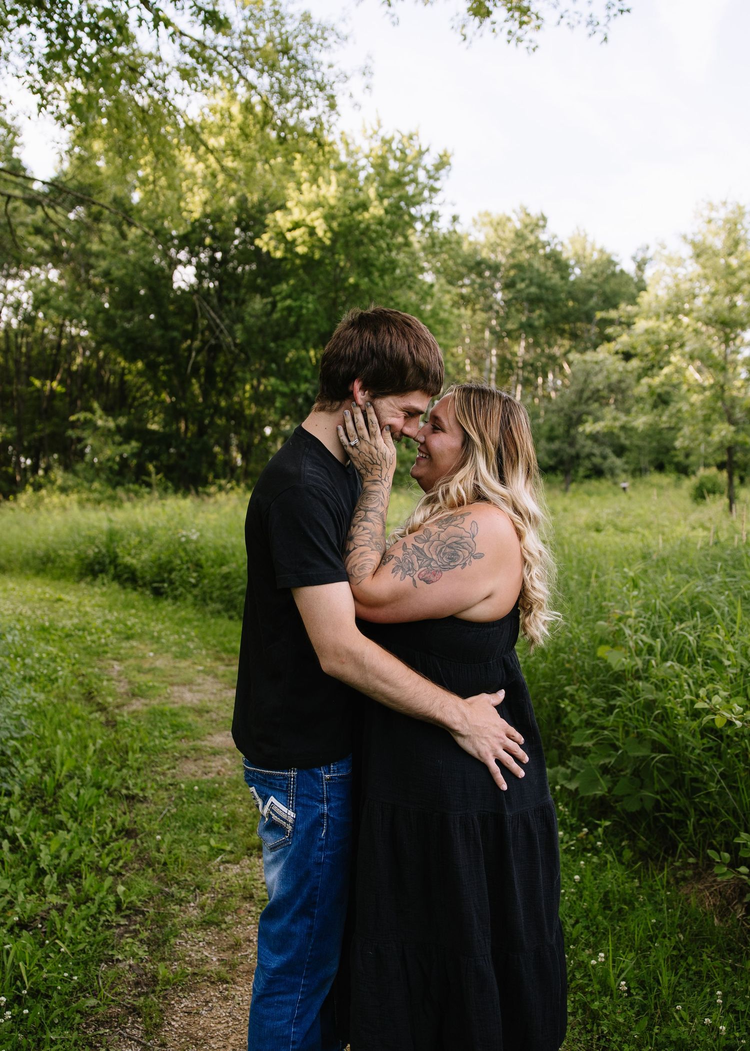 A couple with tattoos shares a romantic moment in a lush green outdoor setting, nearly kissing while embracing.