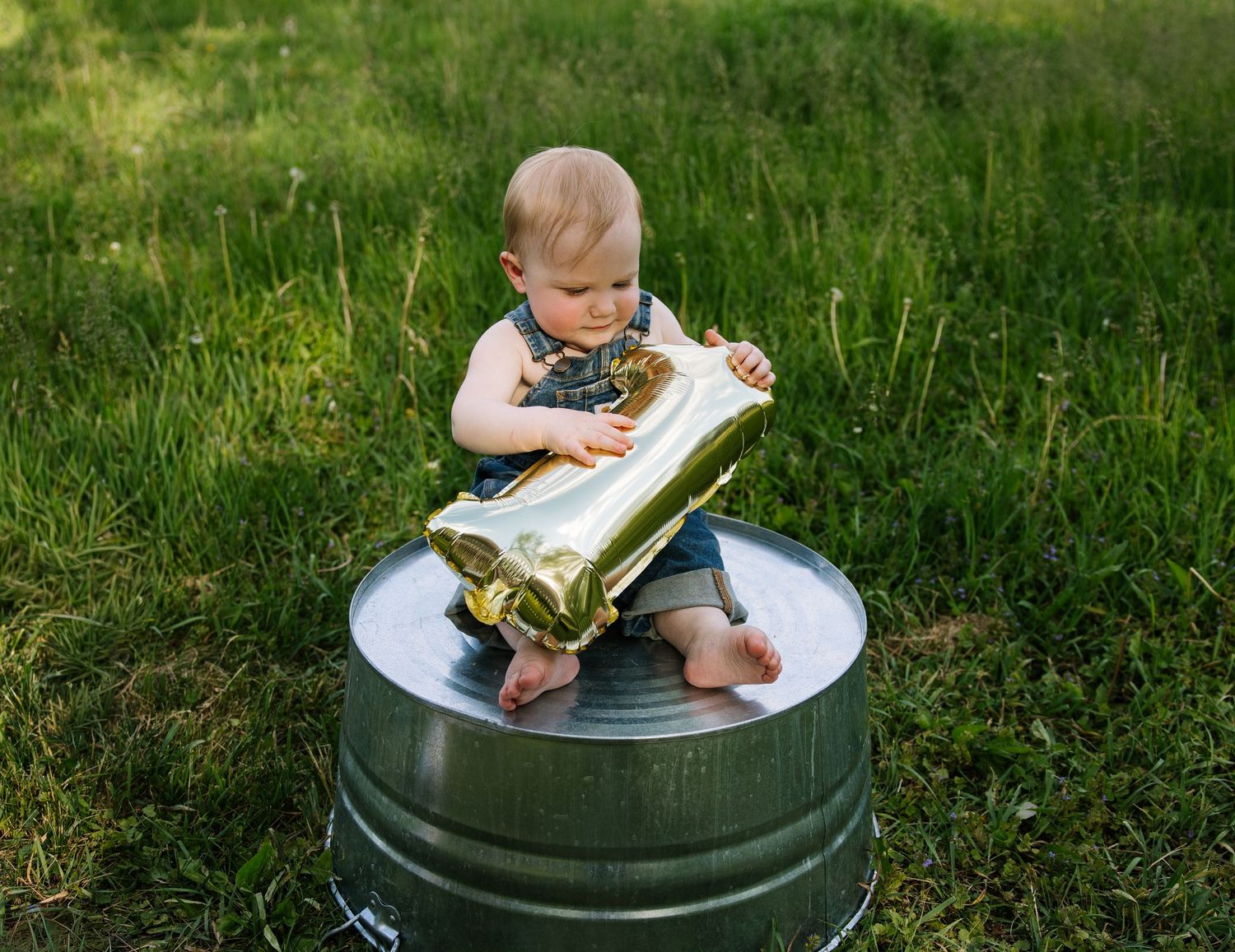 Baby sitting in a metal tub on grass, holding a gold number one balloon, celebrating their first birthday.