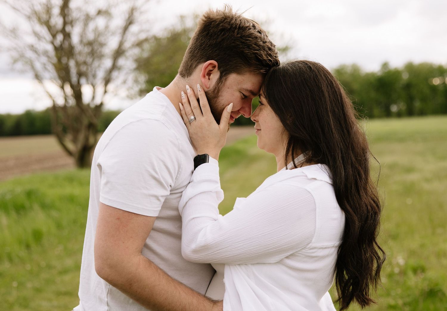 Romantic couple in white embracing forehead-to-forehead in a lush green field, sharing an intimate moment outdoors.