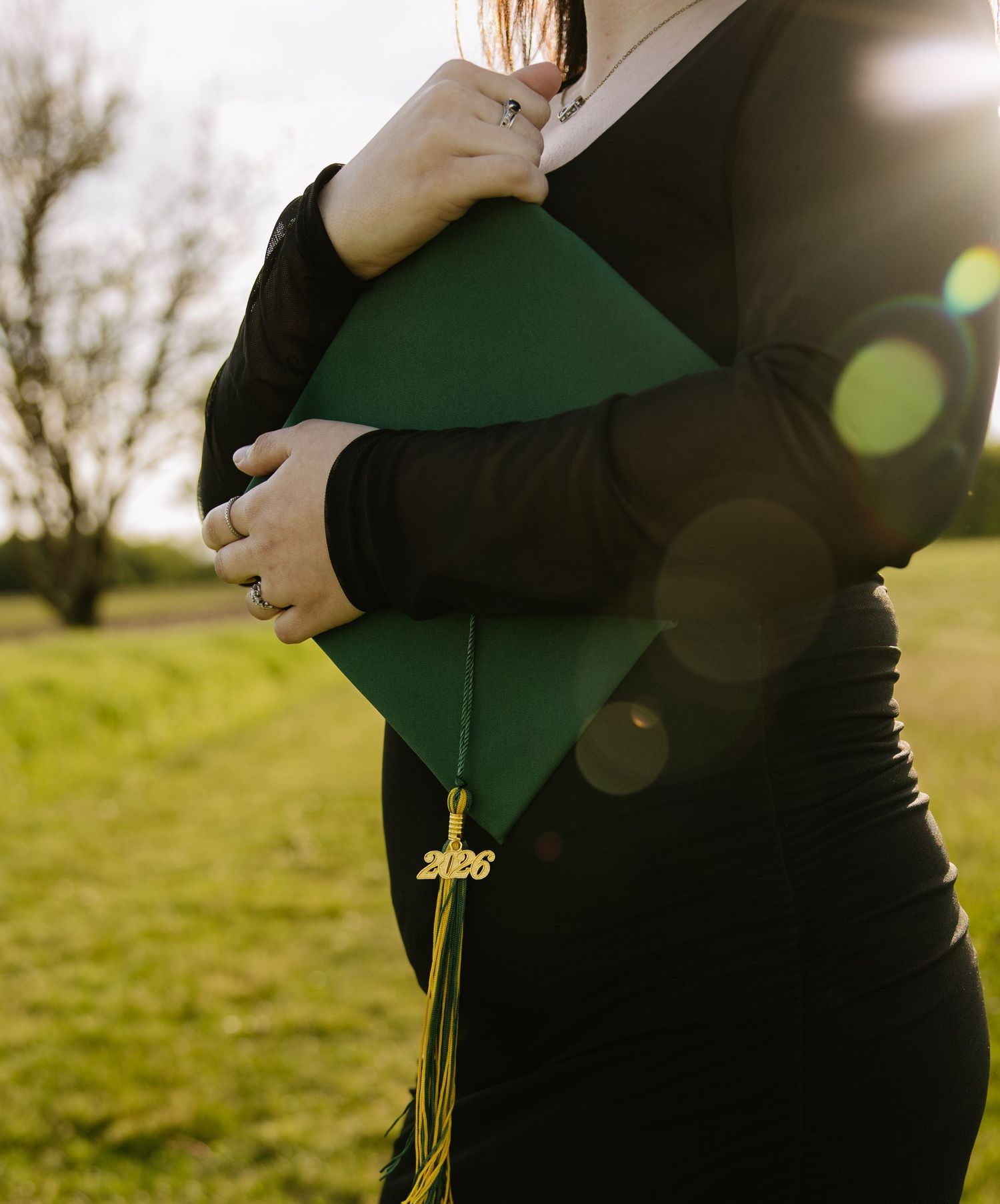 Person in black dress holding green graduation cap with gold 2026 tassel outdoors in sunlit field.