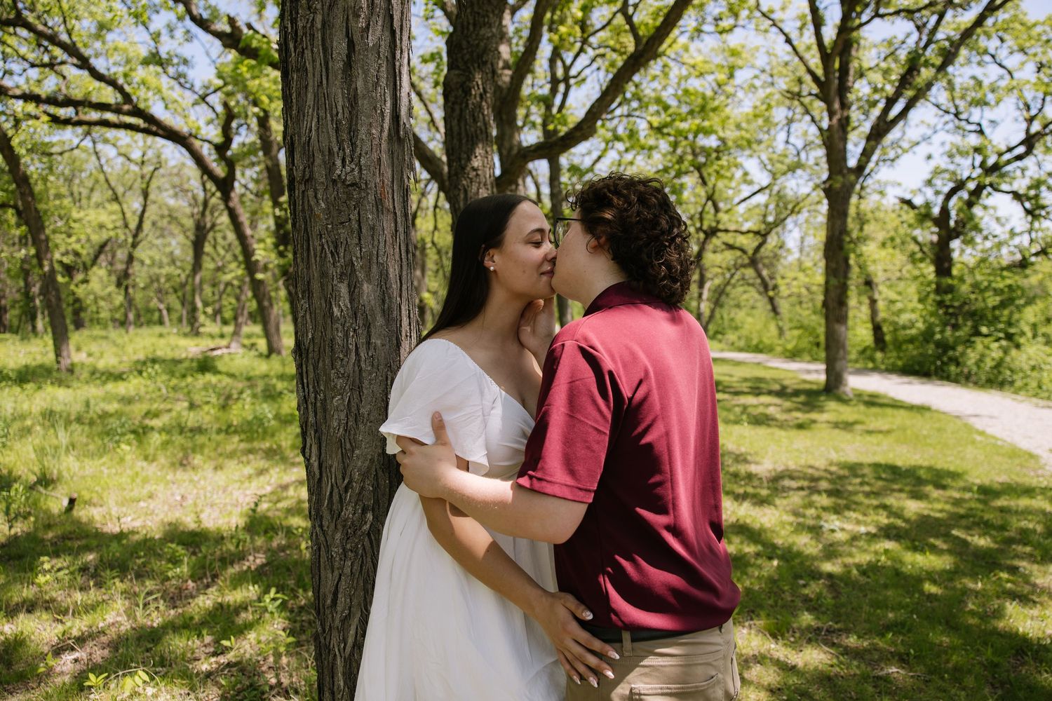 A couple shares a romantic kiss near a tree in a lush green park, with the woman wearing a white dress.
