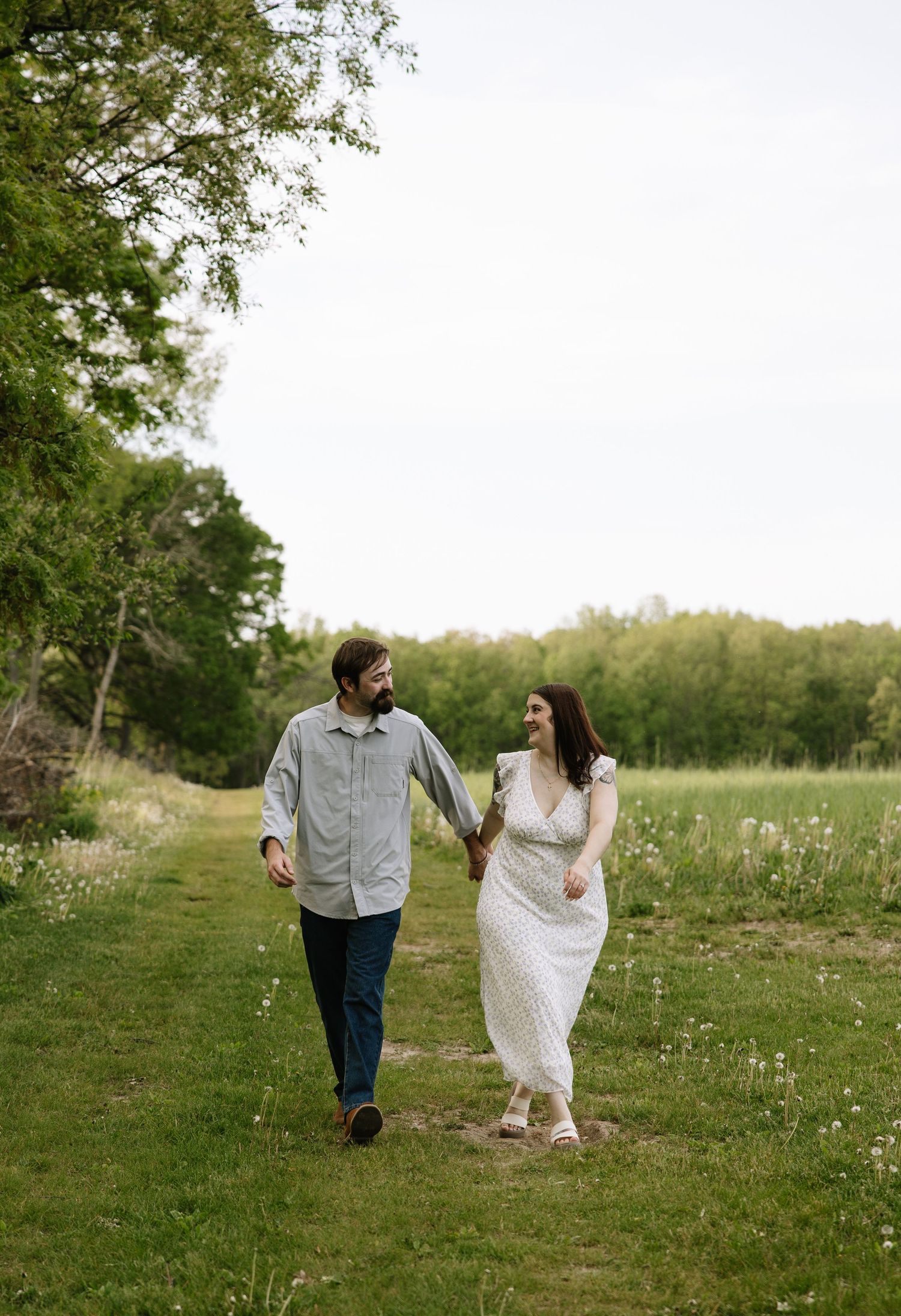 A couple walks hand-in-hand through a lush green meadow path, smiling at each other in a romantic outdoor setting.