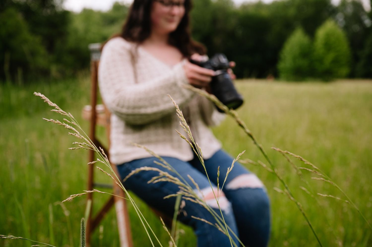 Woman in sweater and jeans sits in field holding camera, with wild grass blades in sharp foreground focus.