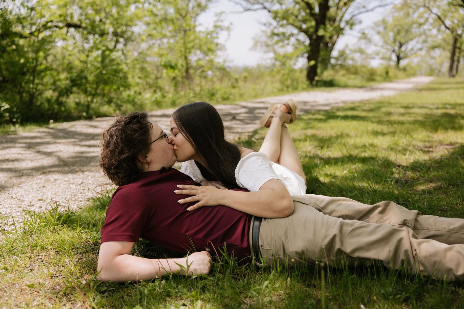 Couple sharing a romantic kiss while lying on green grass in a sunny park setting.