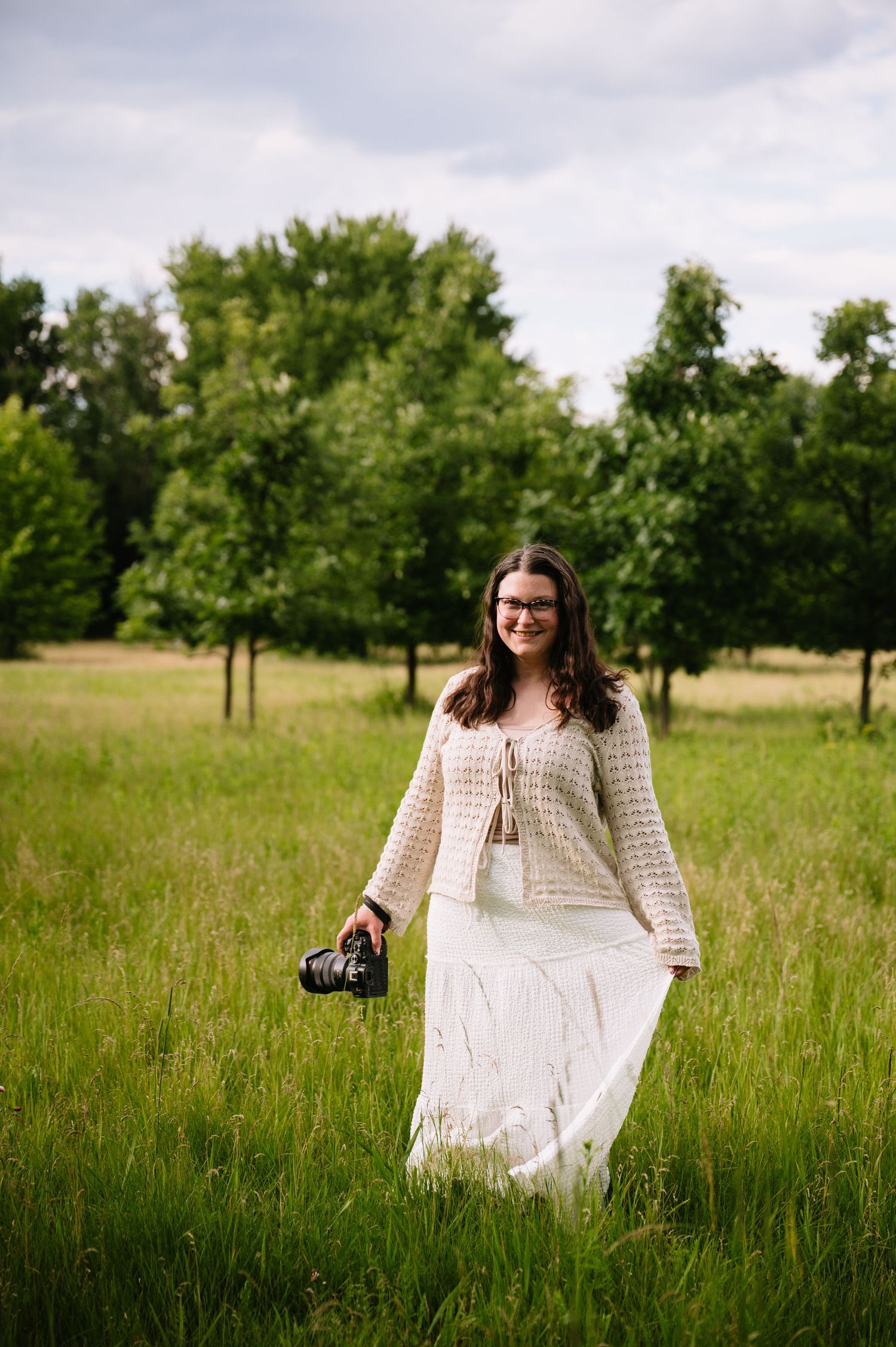 Woman in white skirt and crochet top holds camera while standing in lush green field with trees in background.