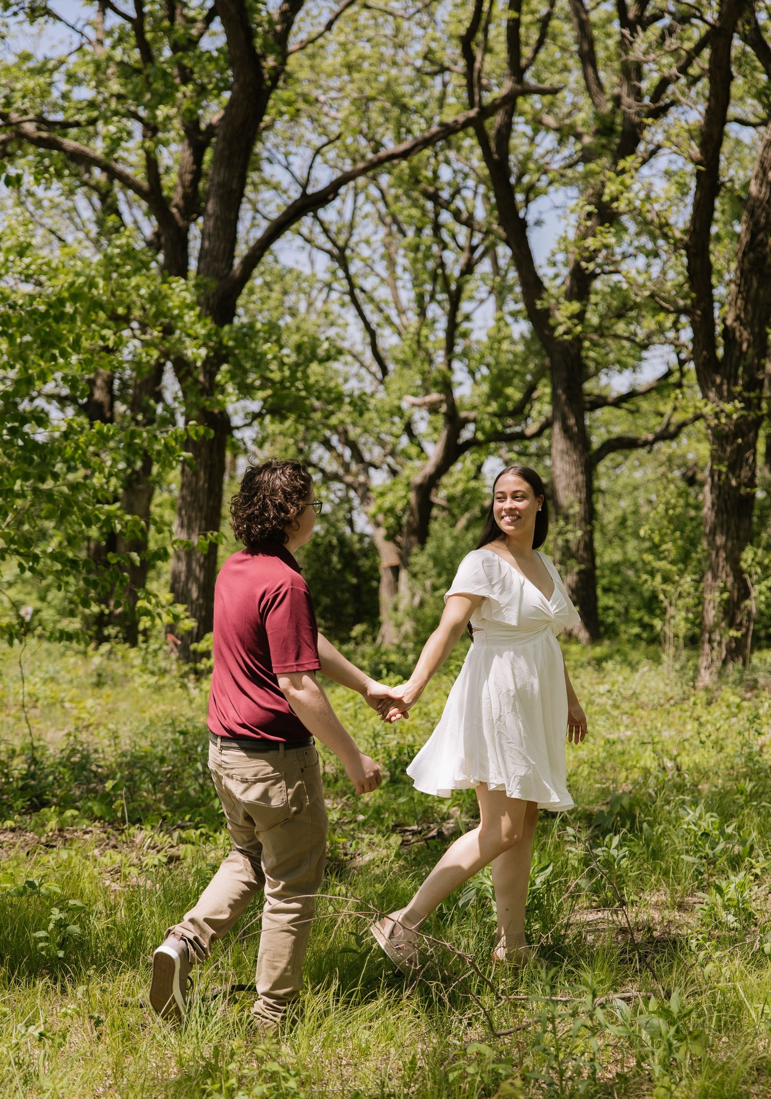 Couple holding hands and walking through a lush green forest clearing on a sunny day.