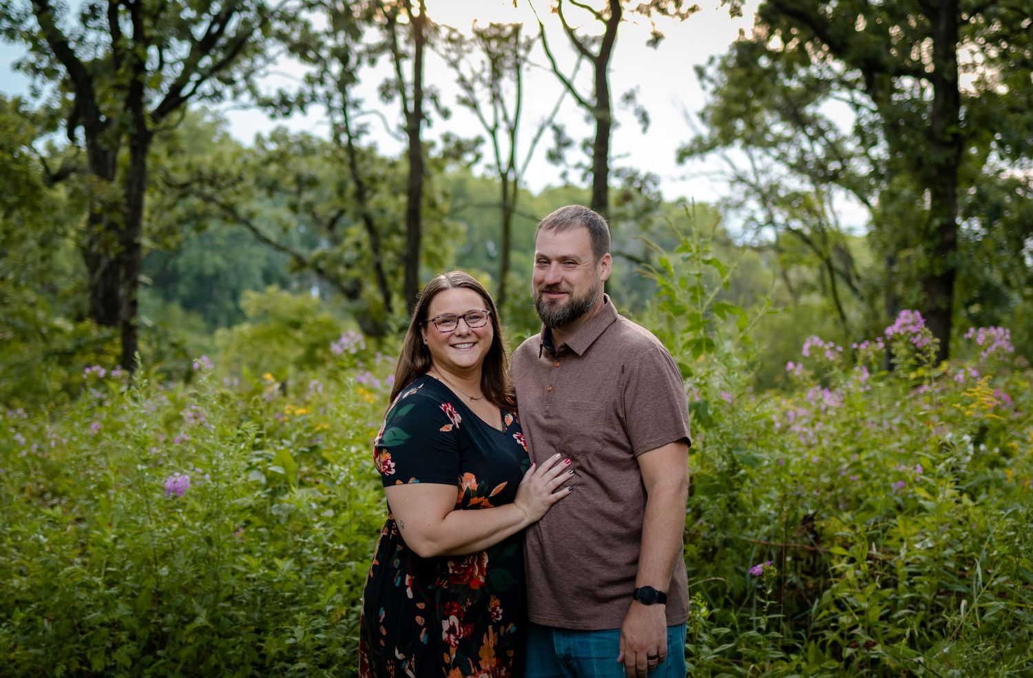 Couple smiling together outdoors surrounded by lush green trees and wildflowers in a natural setting.