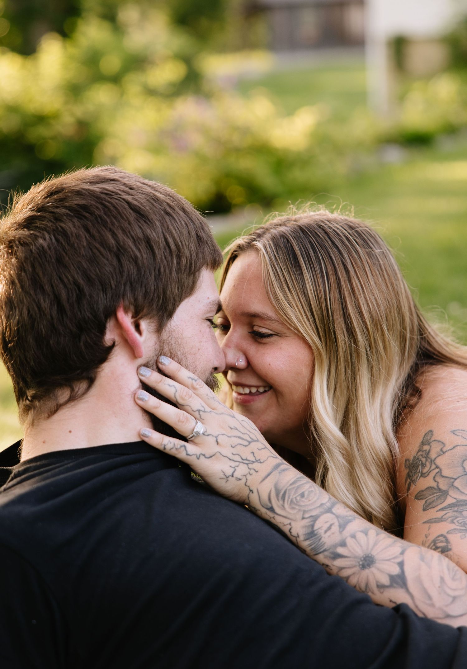 A couple with tattoos shares a romantic moment outdoors, the woman smiling as she holds the man's face, showing her engagement ring.
