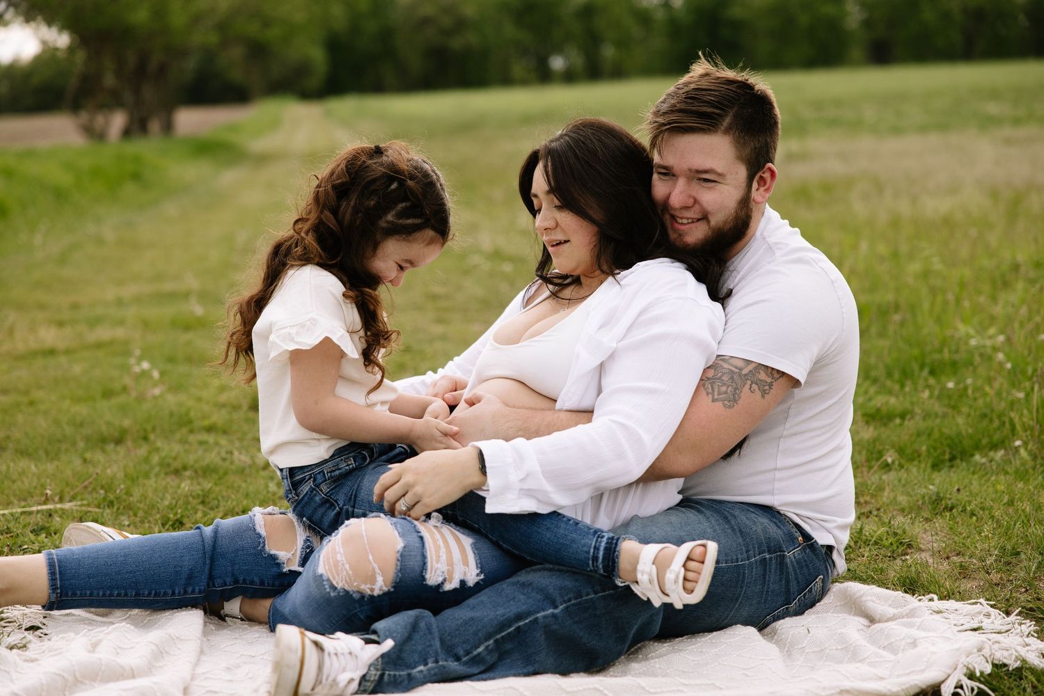 Family maternity photo session outdoors on blanket in green field, parents and daughter touching pregnant belly.