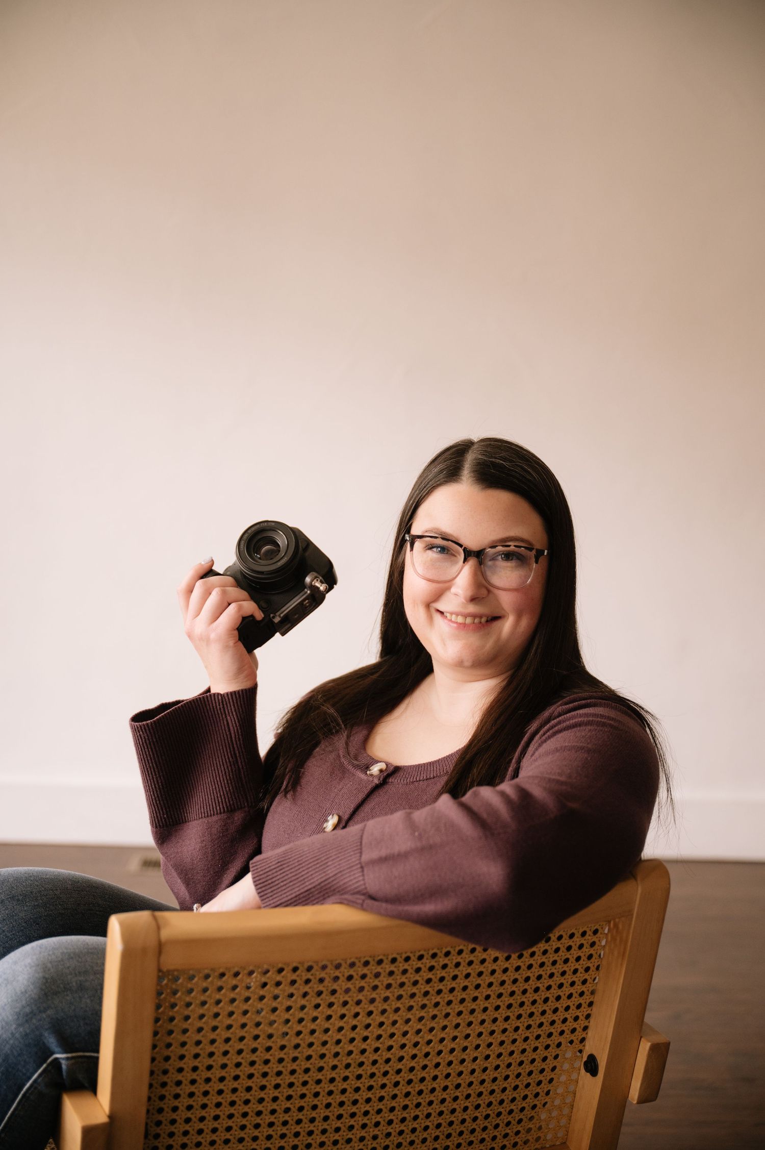 A person in glasses and a brown sweater sits in a wooden cane-backed chair making playful poses.