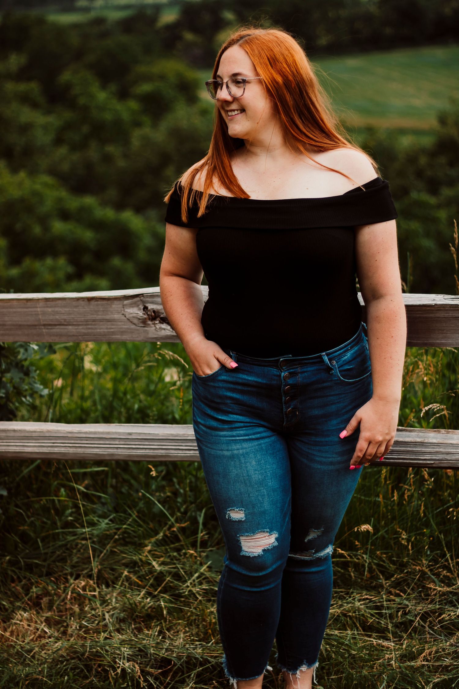 Casual portrait of someone in an off-shoulder black top and ripped jeans leaning against wooden fence.