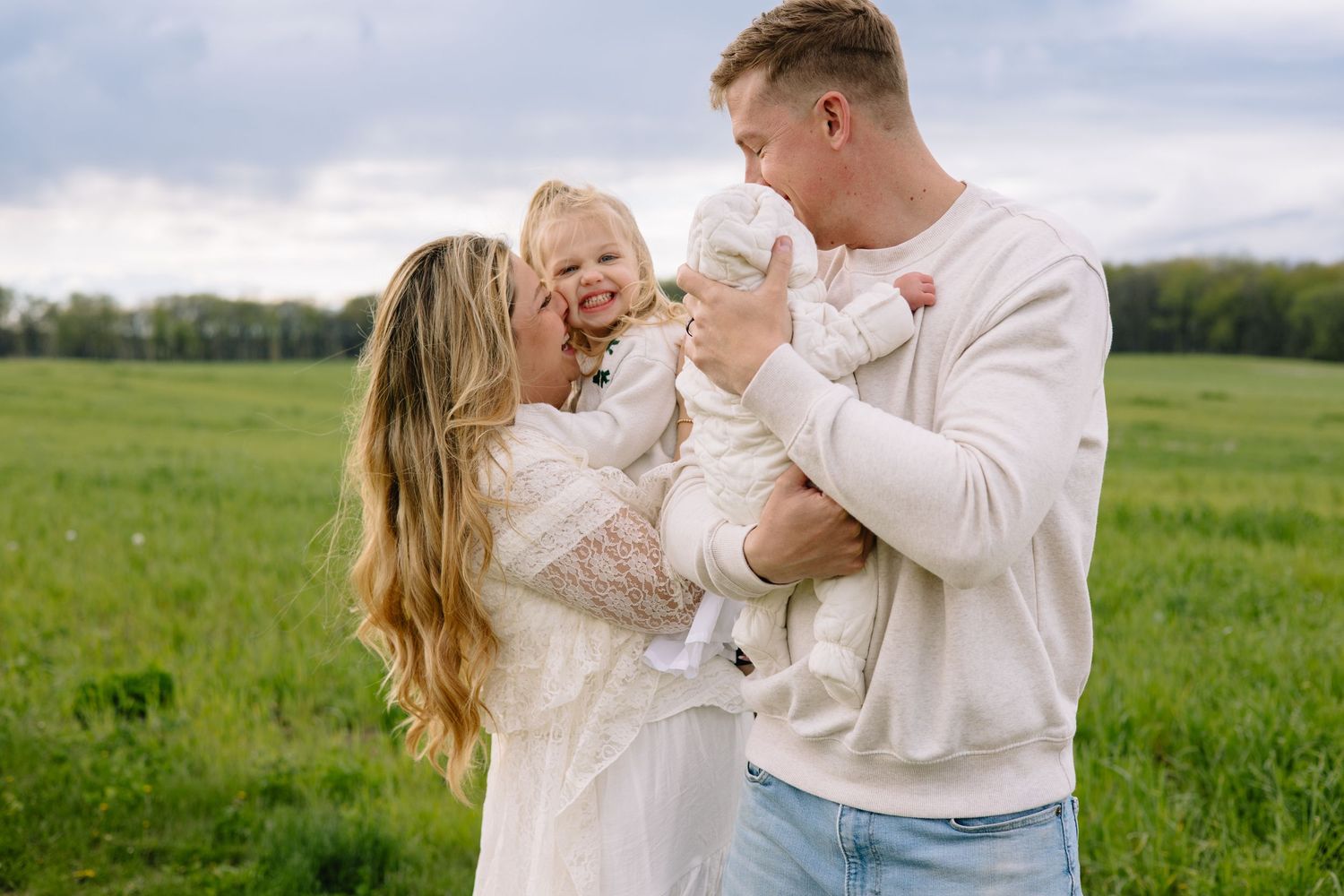Happy family with baby in white outfits posing in green field.