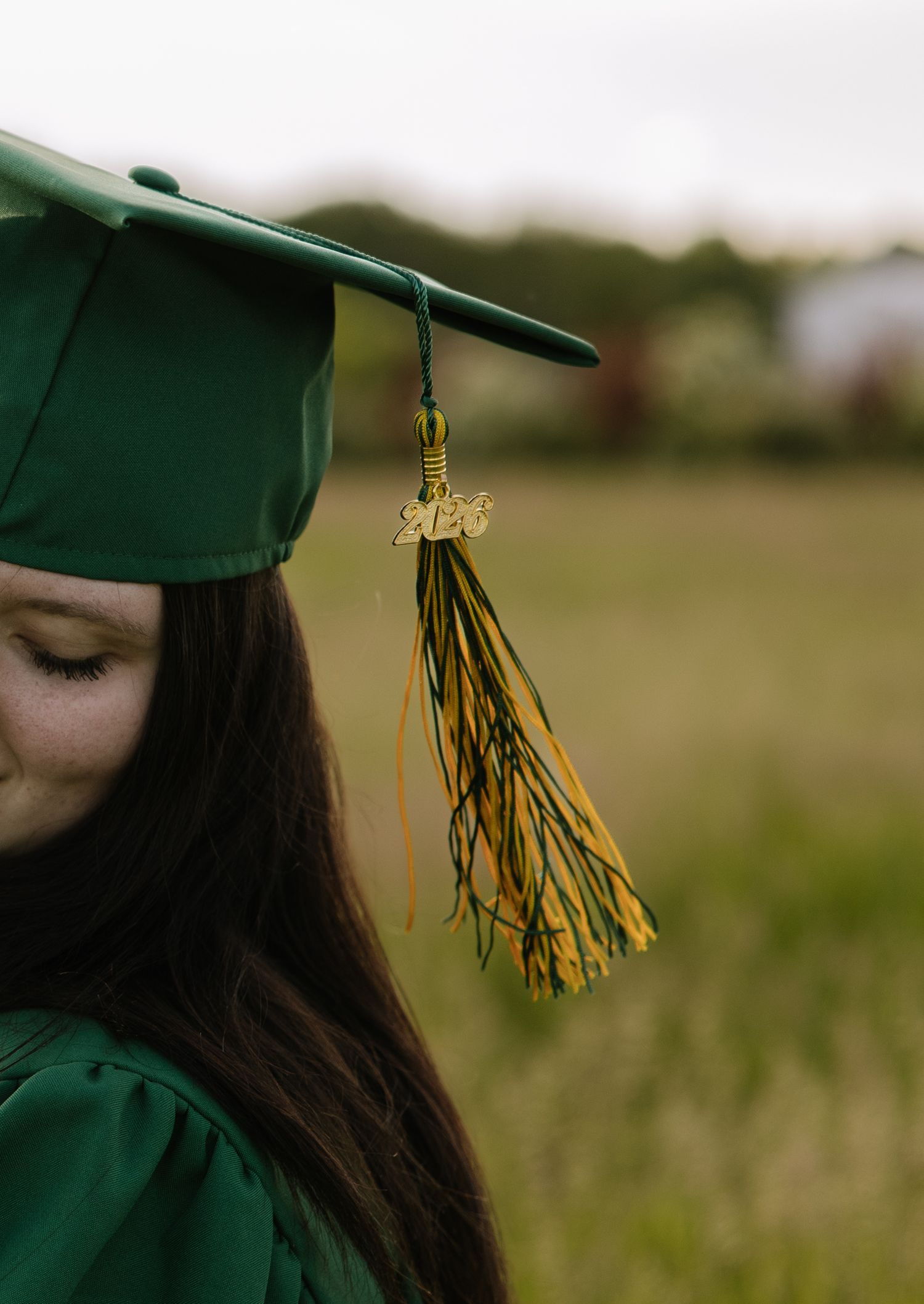 A young woman in a green graduation cap and gown poses outdoors with a gold and green tassel.