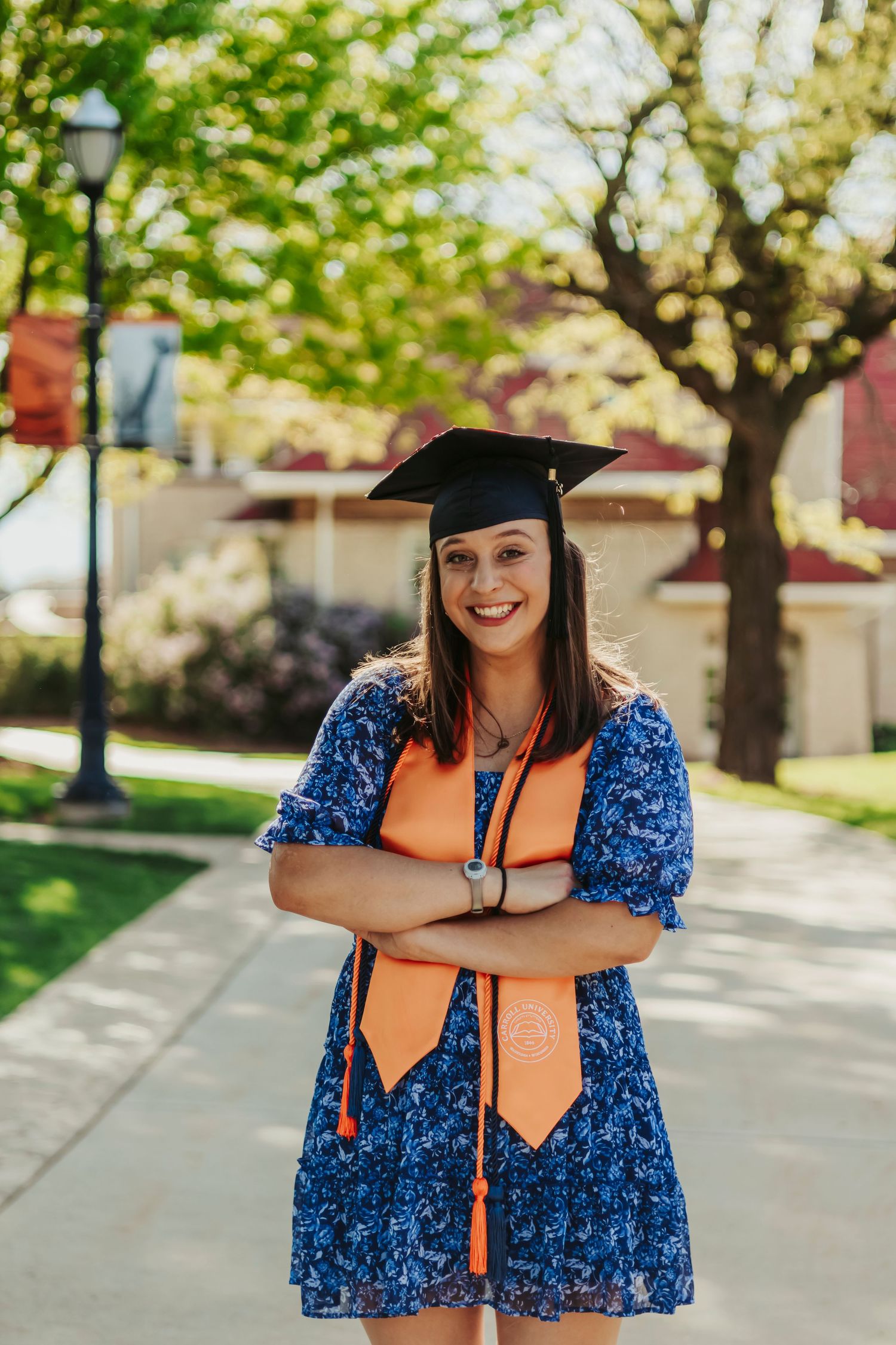 Graduate in cap and gown with blue dress poses confidently on campus walkway.