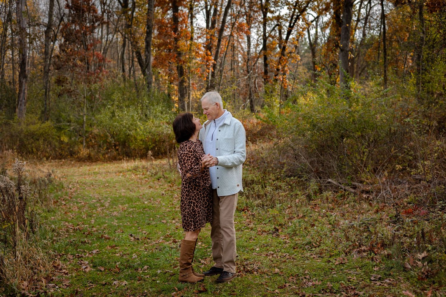 A person in a light blue shirt walks through autumn woods with fallen leaves and bare trees in the background.