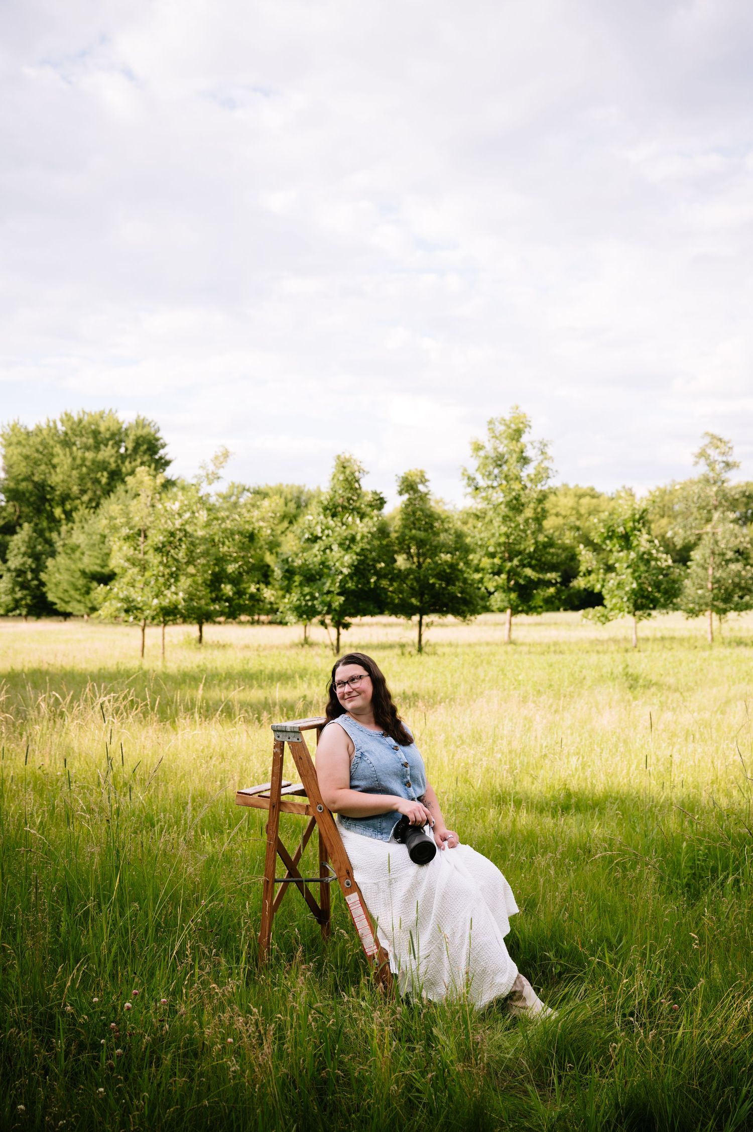 Woman in denim top and white skirt sits on wooden ladder in green meadow holding camera, surrounded by trees.