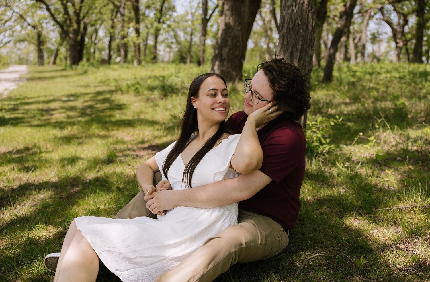 Couple sitting together against a tree in a lush green park, woman in white dress smiling warmly.