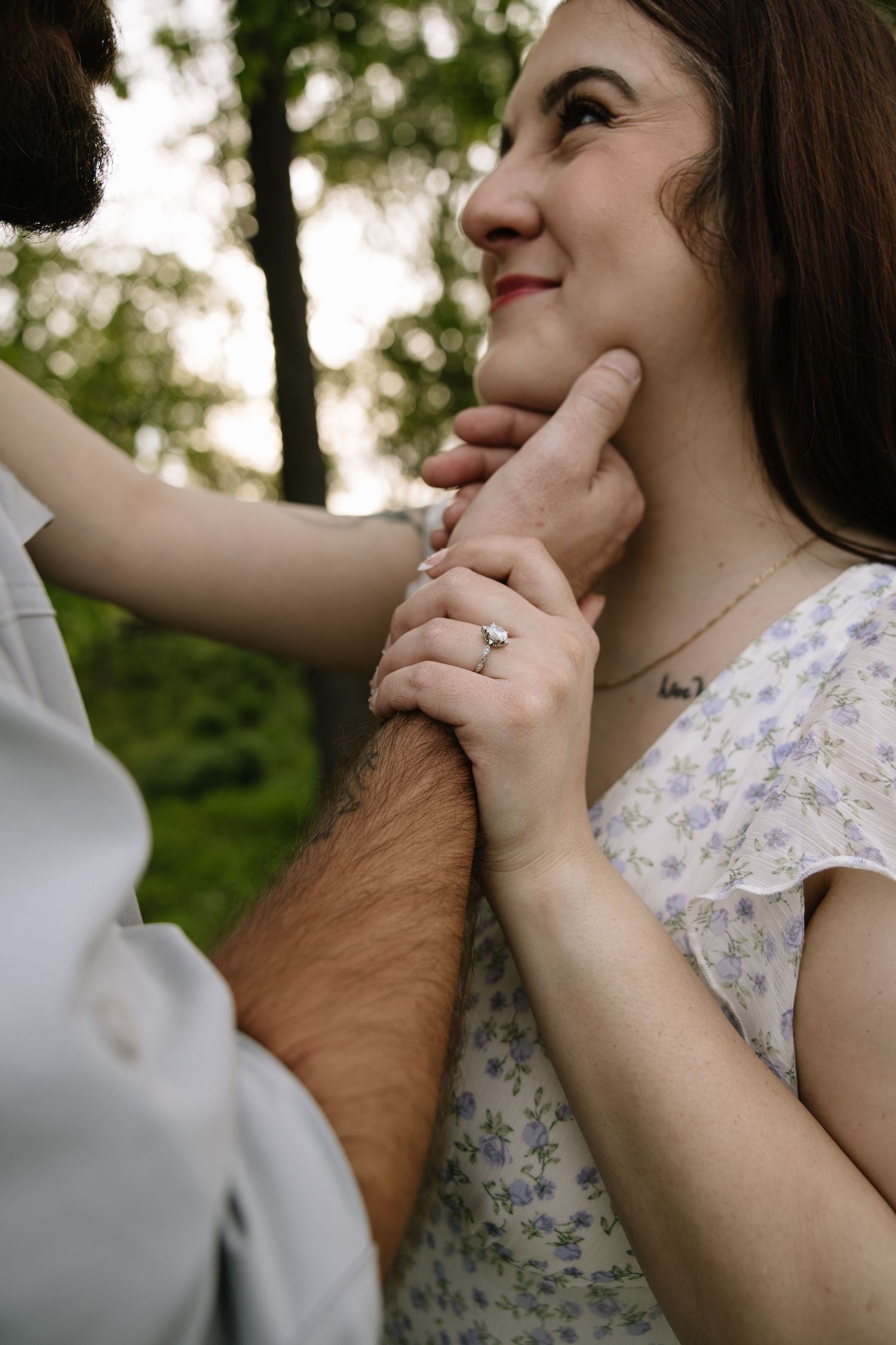 A smiling woman gazes up as her partner gently lifts her chin, showcasing her diamond engagement ring outdoors.