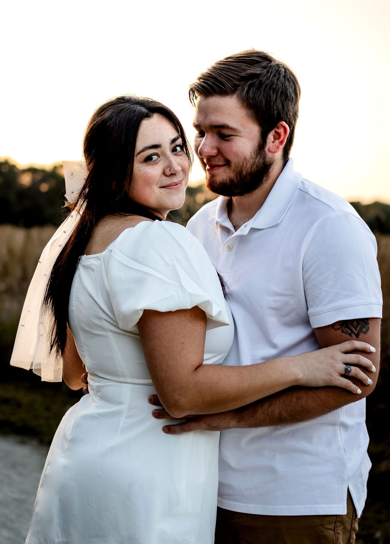 A romantic couple in white outfits embrace during a sunset photoshoot in a natural outdoor setting.