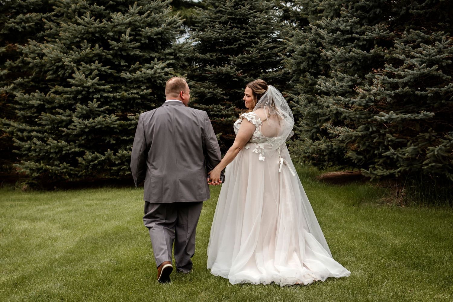 A wedding couple walks hand in hand through a garden path lined with evergreen trees.