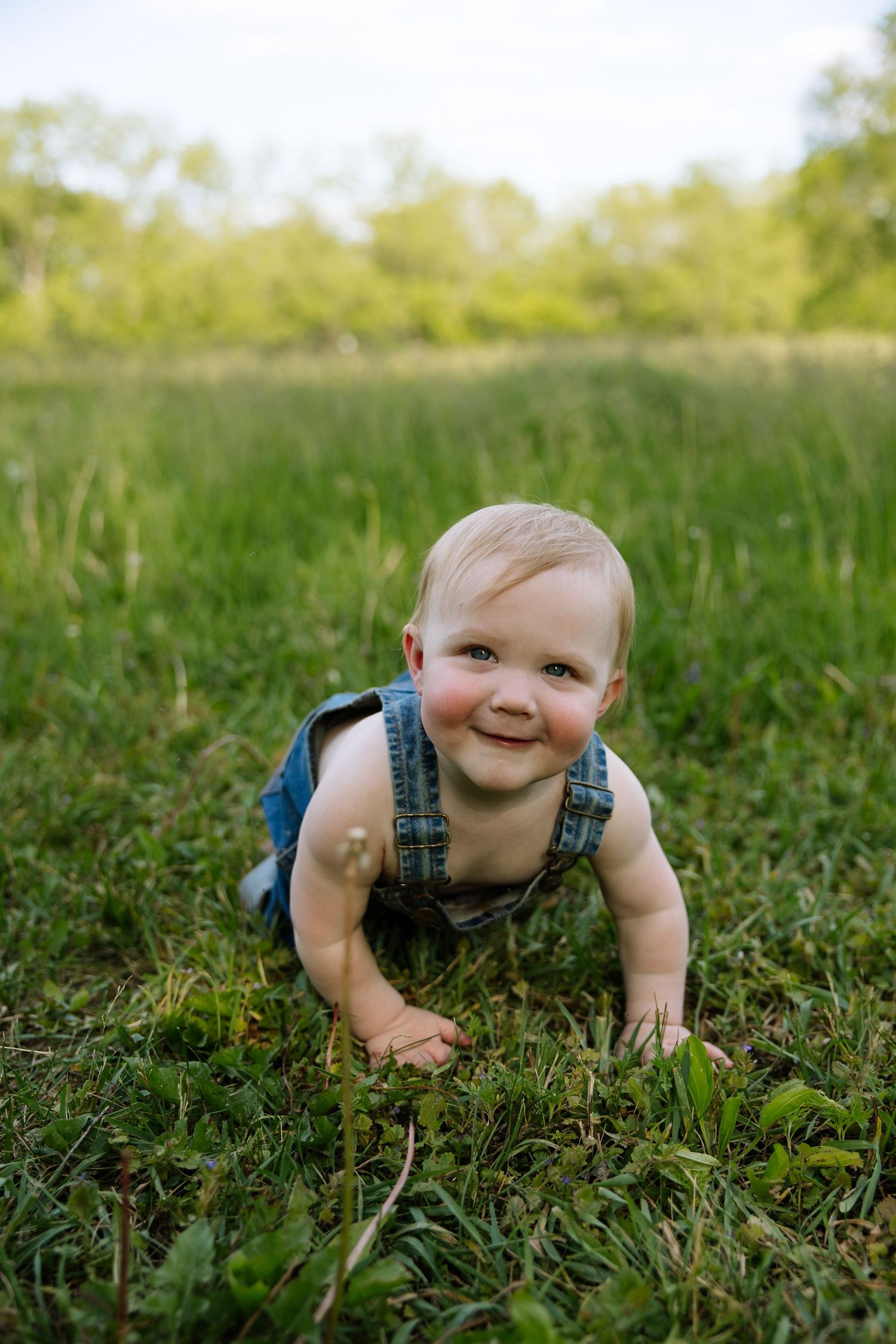 A smiling baby in denim overalls crawling on green grass in an outdoor field on a sunny day.